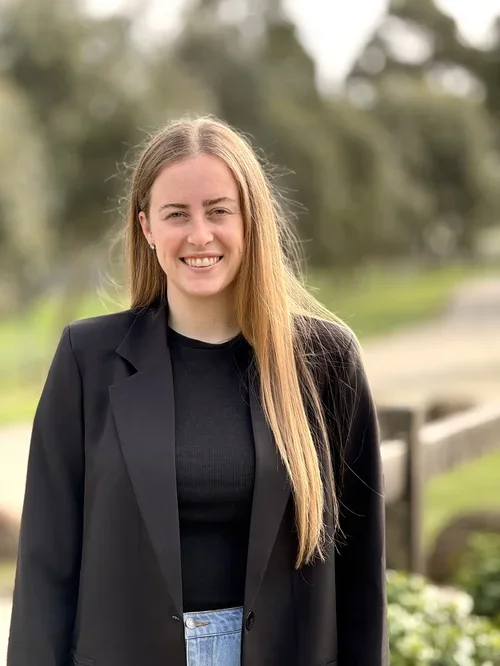 A young speech pathologist who specialises in autism assessments in Bendigo with long blonde hair smiling outdoors, wearing a black blazer over a black top, with trees and a pathway in the background.