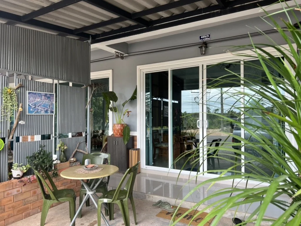 Residential patio with a small round table, four green plastic chairs, potted plants, and a louvered privacy wall, overlooking a glass-door entrance to the house.