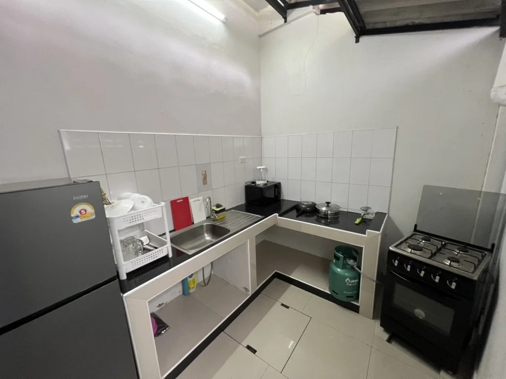 Small kitchen with white tiled wall, white countertop, gray refrigerator, black stove, microwave, and various kitchen items.
