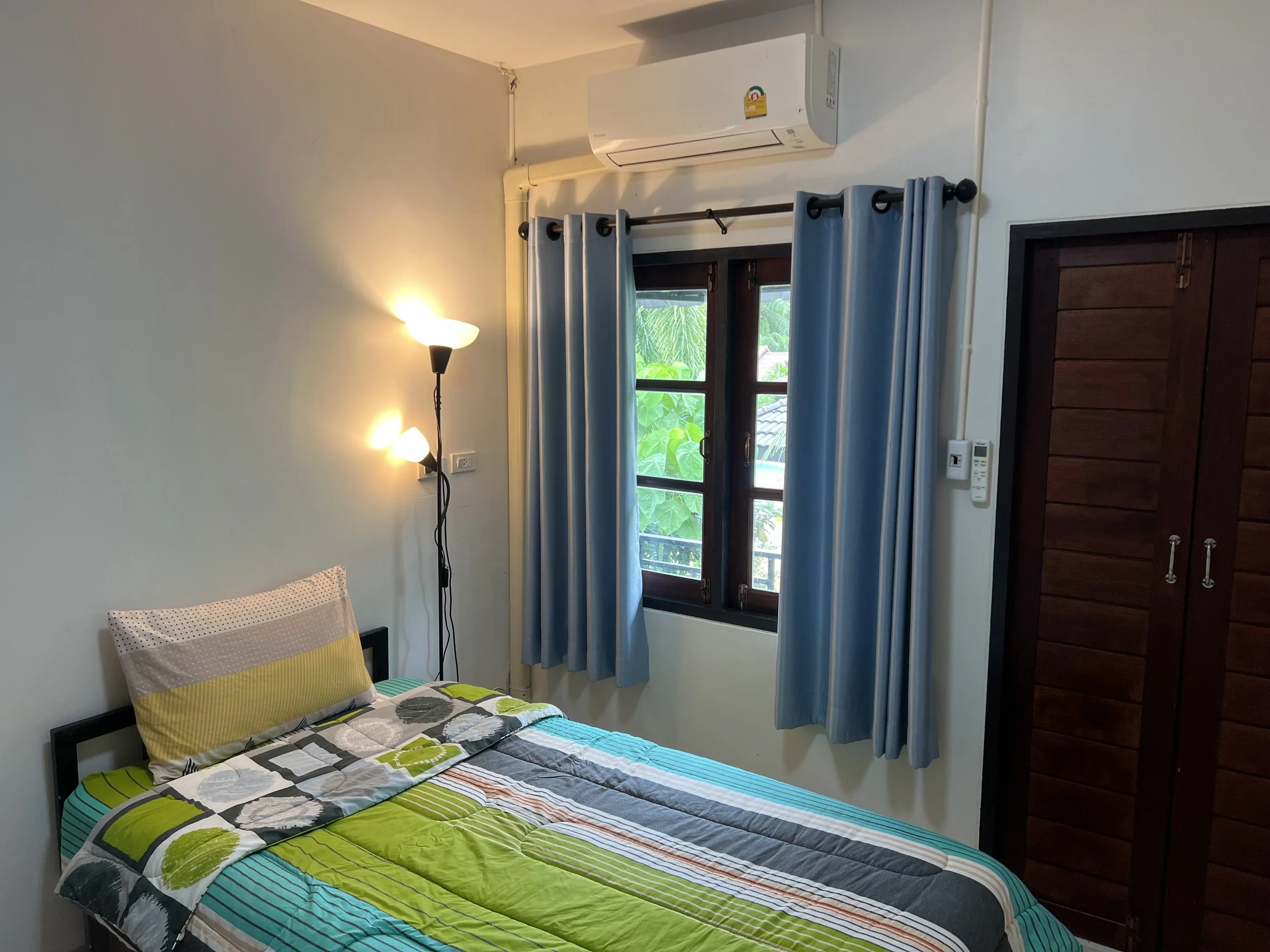 A bedroom window with blue curtains, a bed with a colorful striped comforter and pillows, a wall-mounted lamp, an air conditioning unit above the window, a wooden wardrobe, and green foliage outside the window.