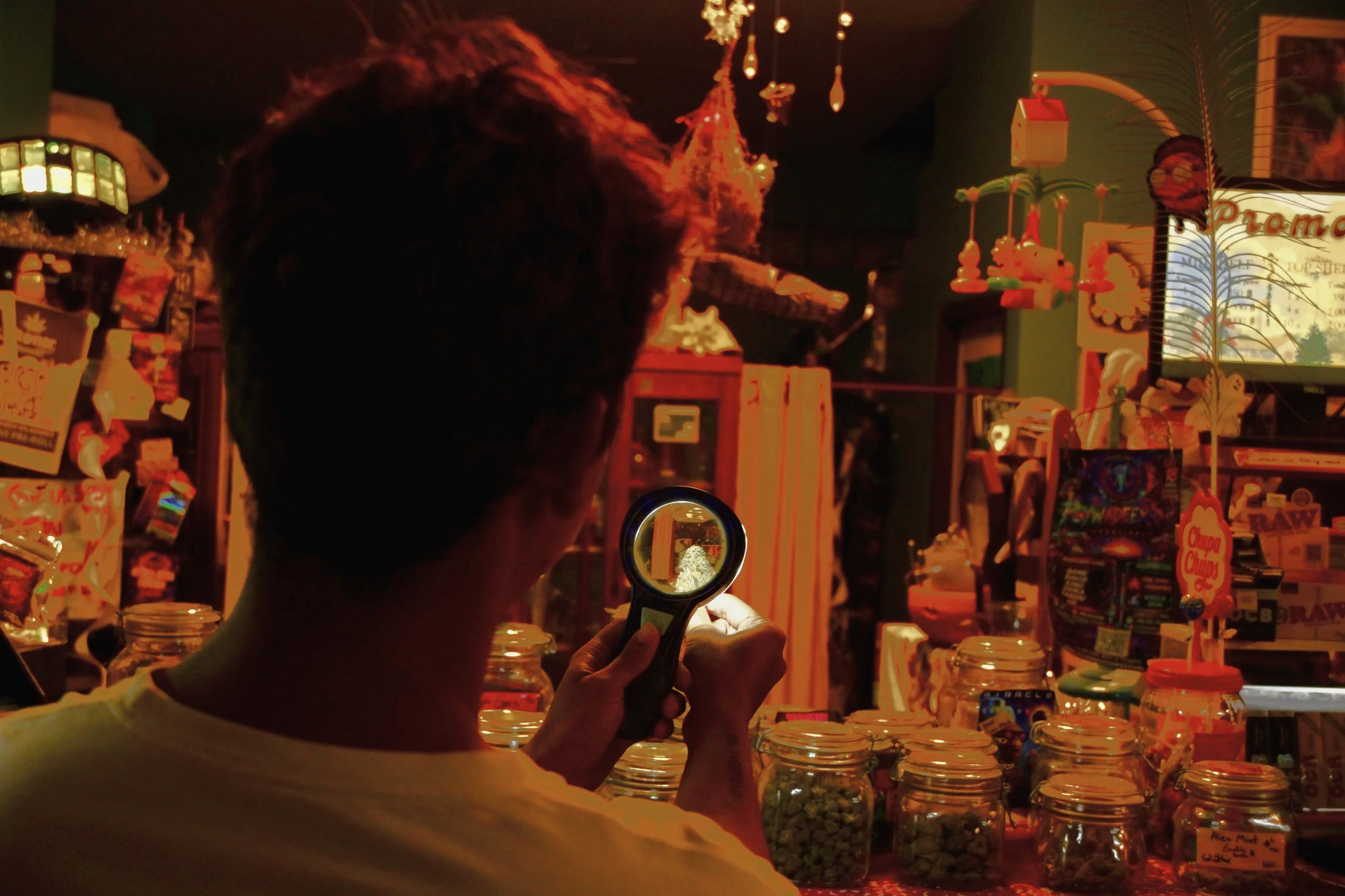 Person holding a flashlight in a store aisle with jars and colorful decorations, including hanging toys and signs, illuminated by a dim, reddish light.