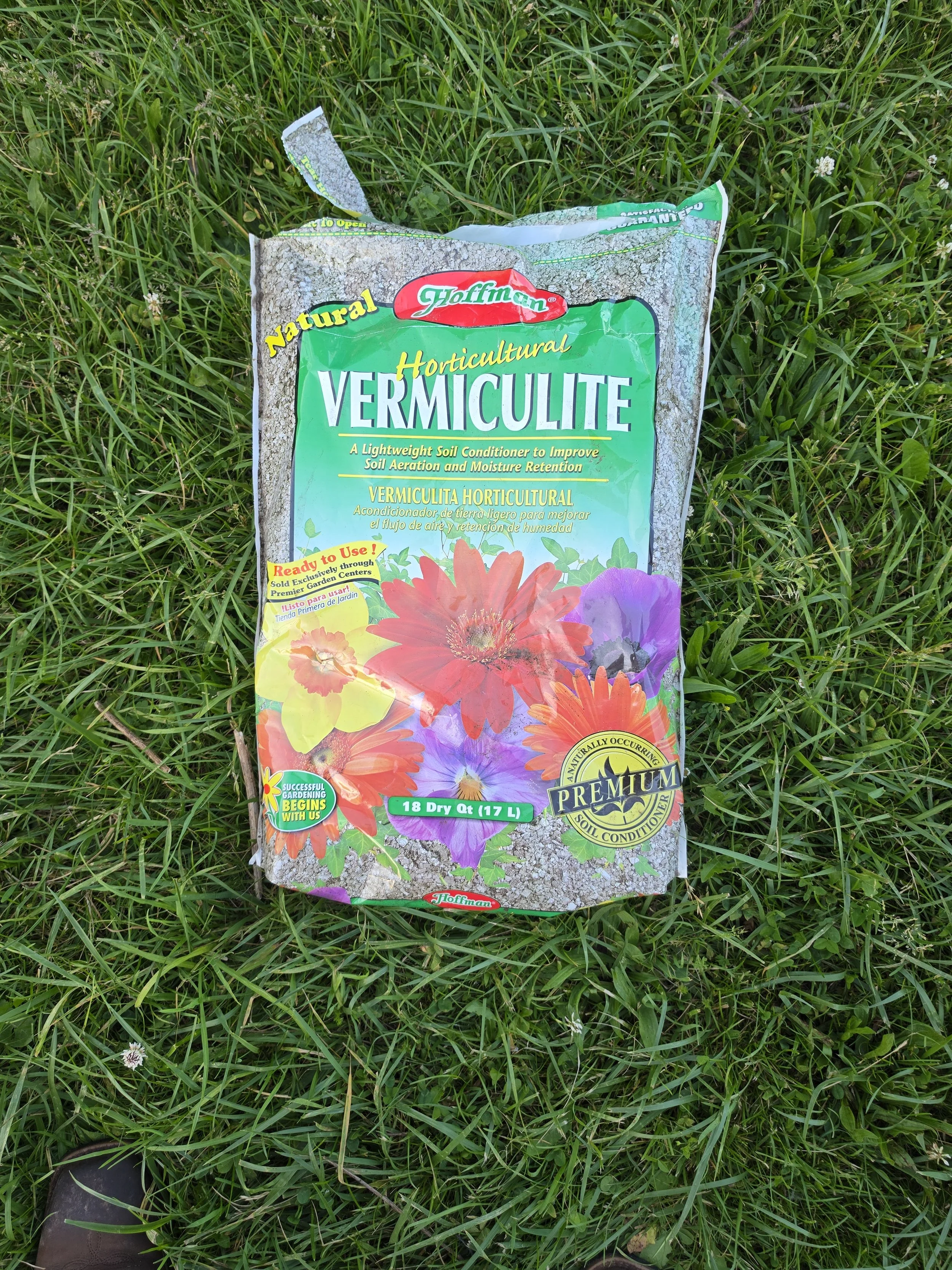 A bag of Hoffman horticultural vermiculite lying on green grass, with small white flowers and part of a person's shoe visible in the lower left corner.