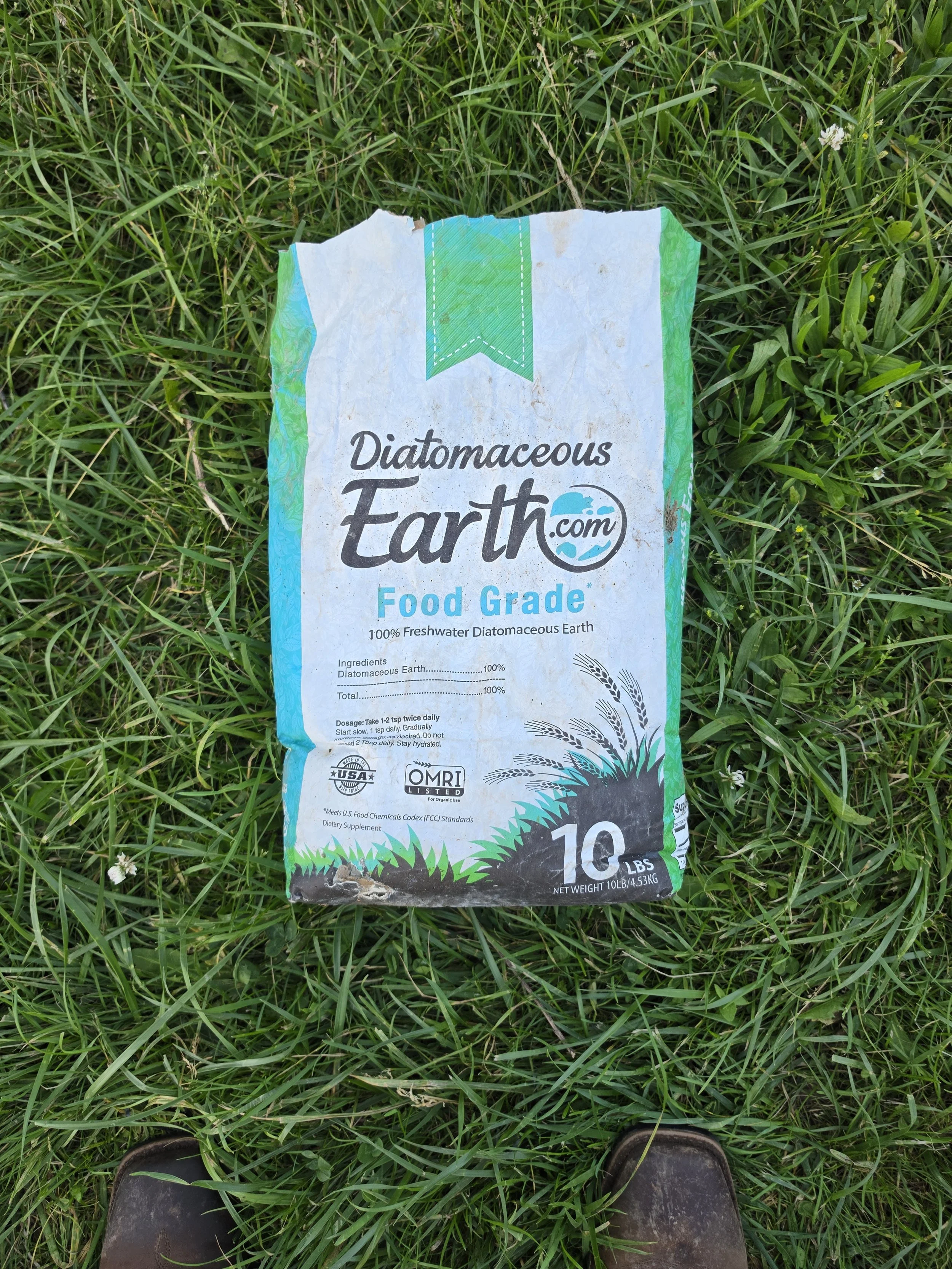 A bag of diatomaceous earth lying on green grass, with part of a person's black shoes visible at the bottom of the image.