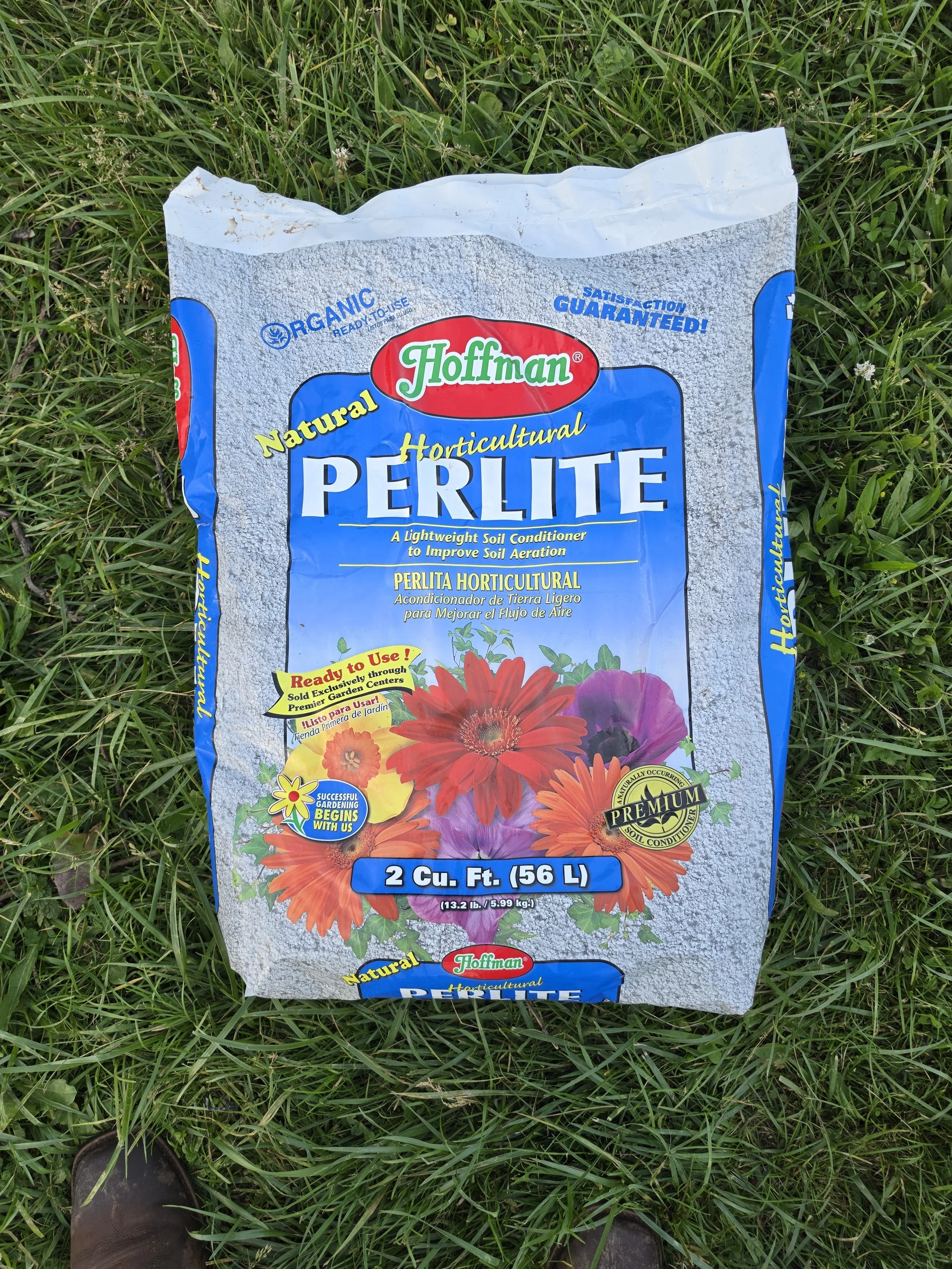 A bag of Hoffman horticultural perlite for soil conditioning, lying on grass with a person wearing brown shoes visible at the bottom of the image.