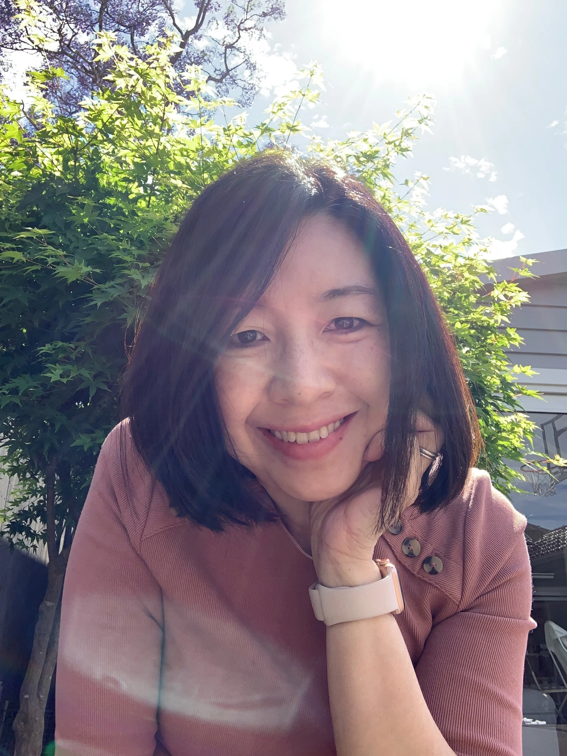A woman smiling outdoors with sunlight shining from behind, green leafy trees in the background, wearing a pinkish-brown top and a white watch.