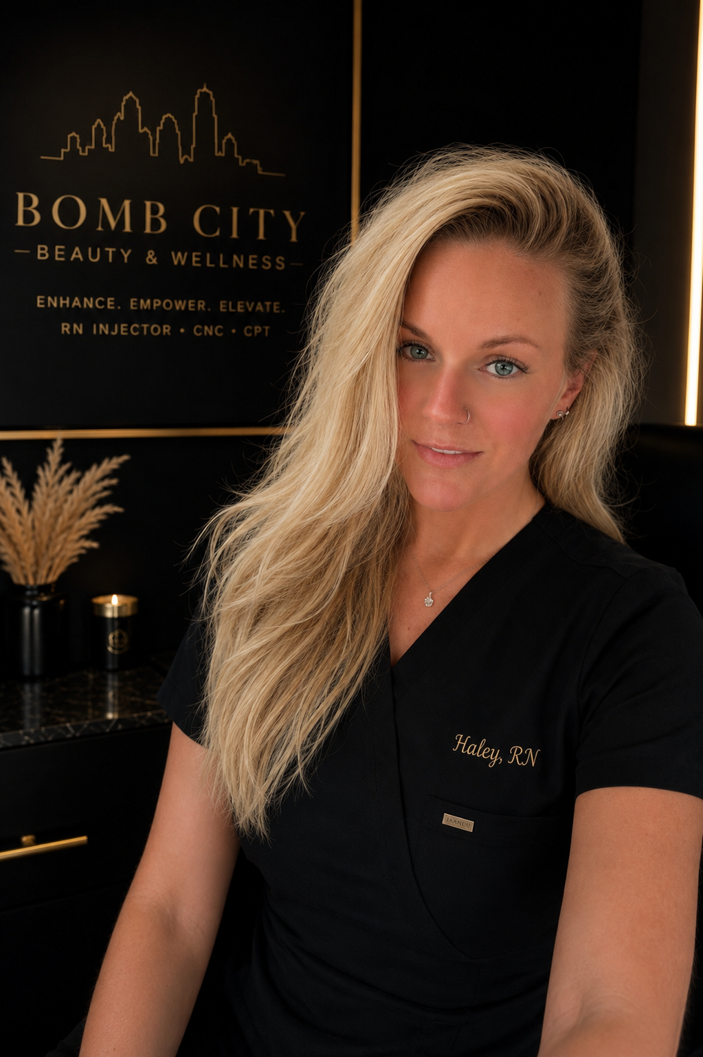 A blonde woman with blue eyes, wearing a black uniform with a name tag that says 'Haley RN'. She is sitting in a stylish, modern spa or wellness clinic with black and gold decor. Behind her on the wall is a sign that reads 'Bomb City Beauty & Wellness' and features an outline of a city skyline.