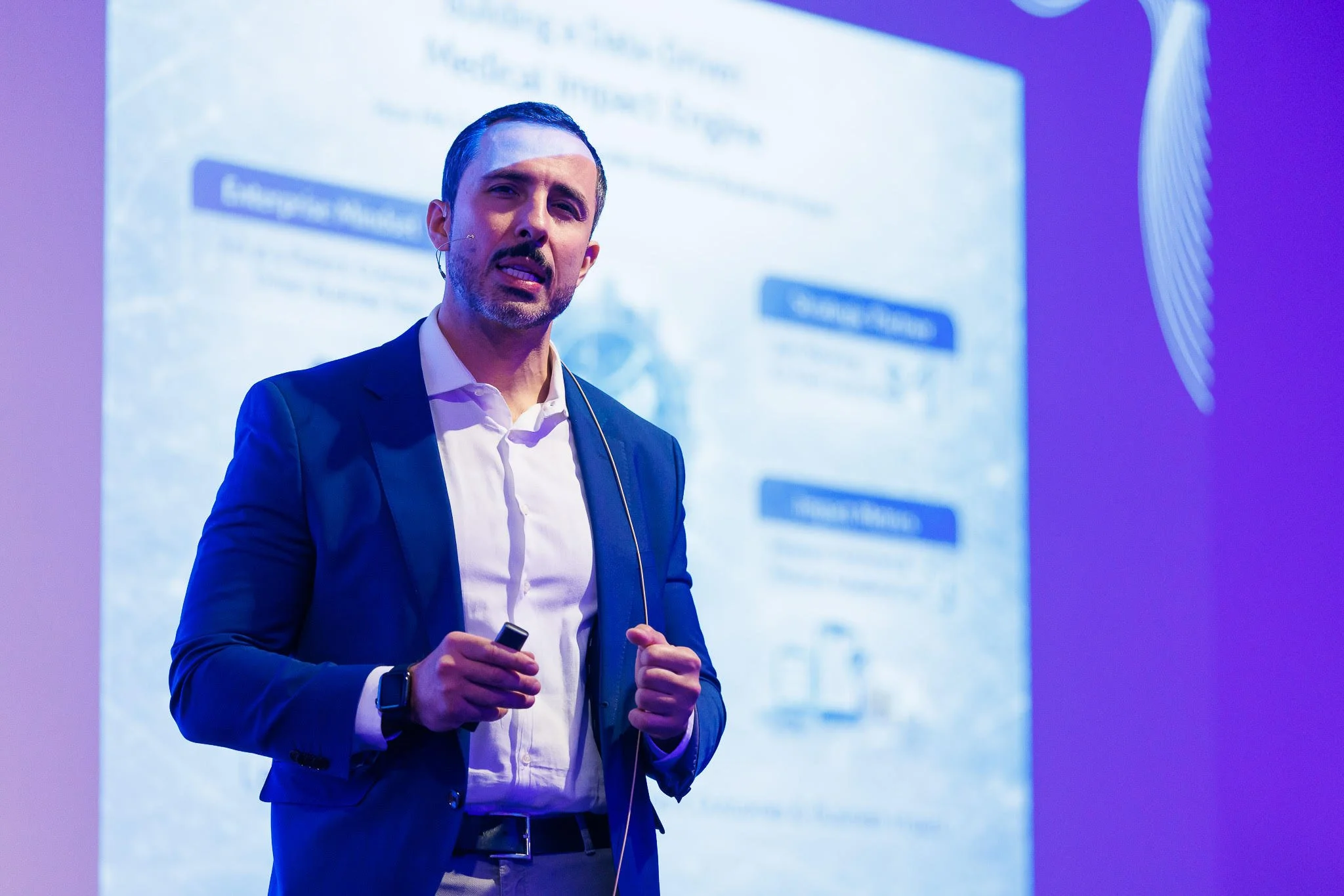 A man in a suit giving a presentation, standing in front of a screen with blurred text, holding a remote in his right hand, with blue and purple lighting.