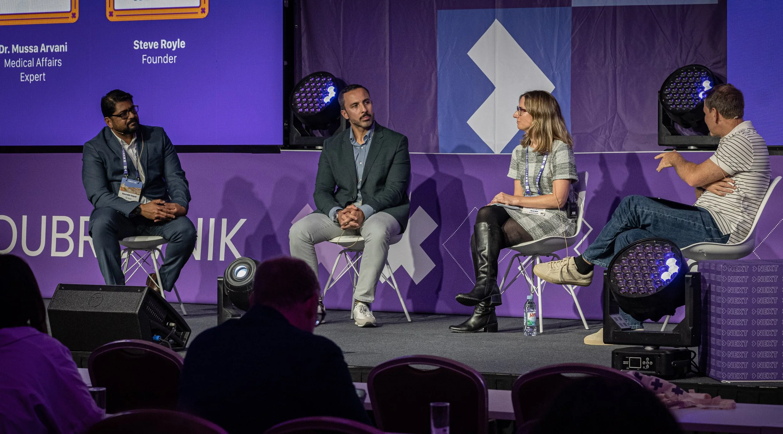 Four people sitting on stage at a conference or panel discussion, with a purple backdrop and large screens displaying text and graphics.