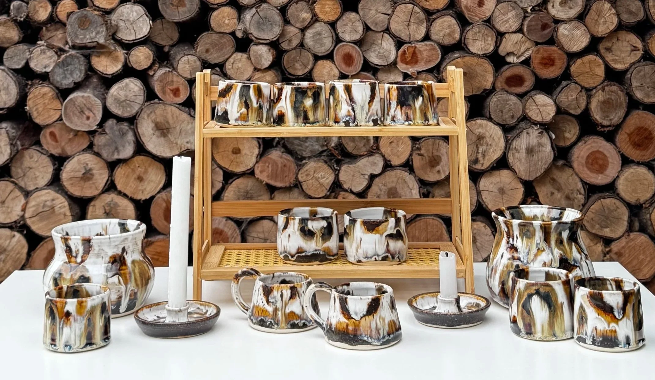A display of marbled ceramic cups, teapots, and bowls arranged on a white table, with a backdrop of stacked logs.