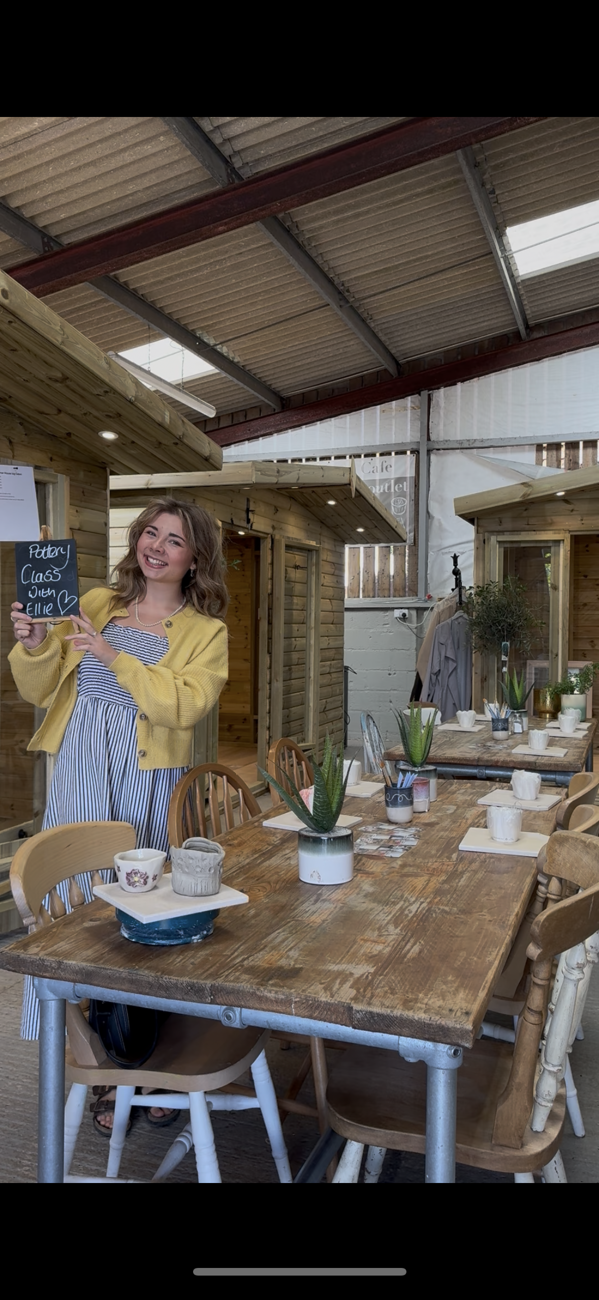 A smiling woman holding a small blackboard sign at a rustic indoor dining area with wooden tables and chairs, decorated with potted plants.