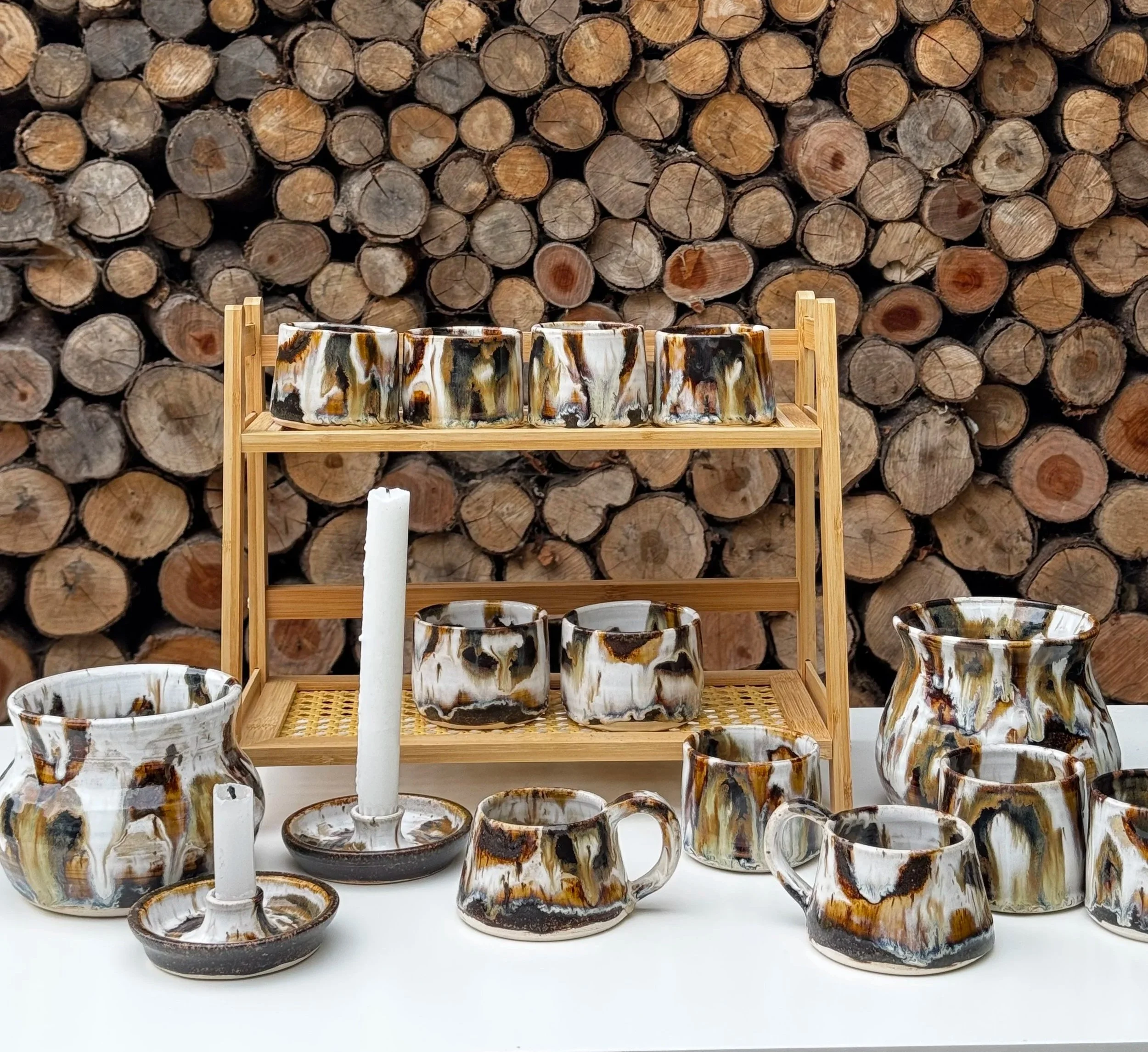 Collection of ceramic vessels with brown, black, and white swirling patterns, arranged on and below a wooden display stand with a backdrop of stacked firewood.