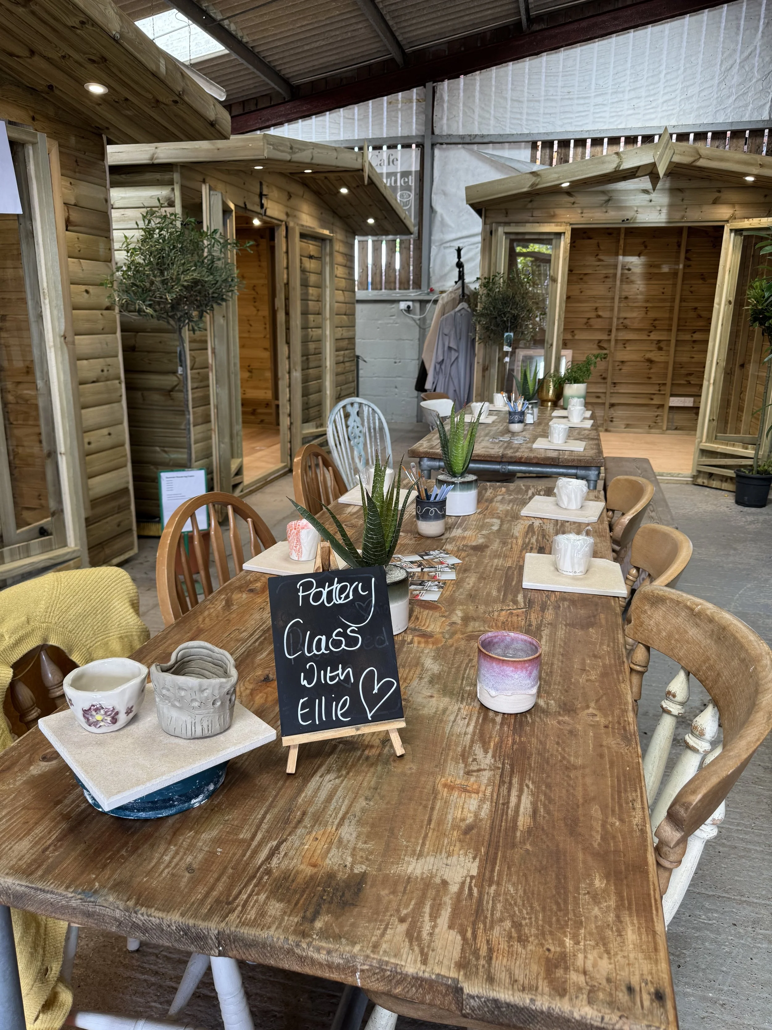 A wooden table displays various handmade pottery items, with a small chalkboard sign reading 'Pottery Class with Ellie' and heart. The background shows a rustic indoor setting with small wooden cabins, potted plants, and tables.