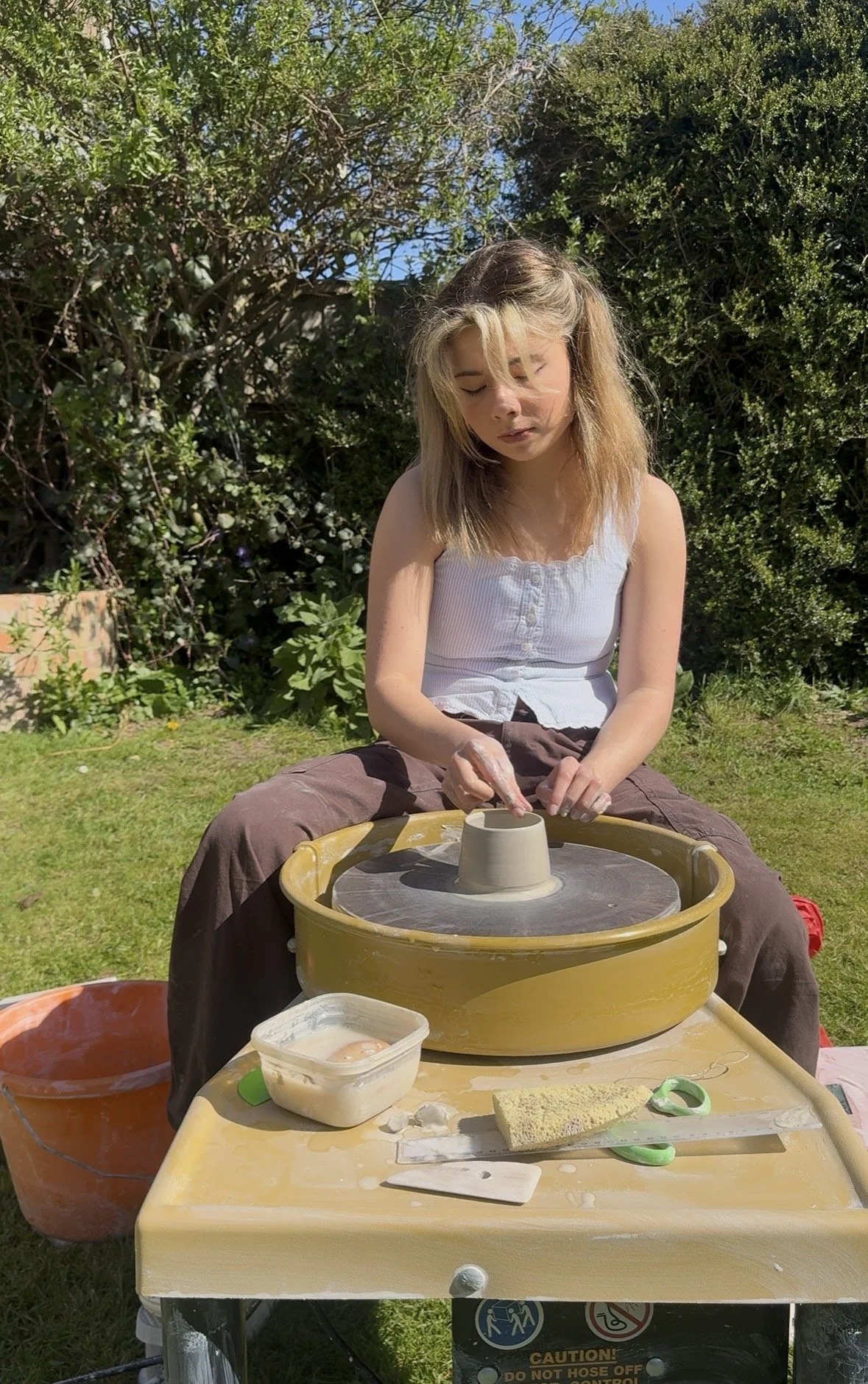 A young woman sitting outdoors at a pottery wheel, shaping clay into a vessel. She is wearing a white sleeveless top and brown pants, with greenery and bushes in the background. There are pottery tools and clay on the table in front of her.