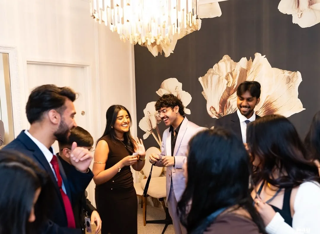 Group of people at a social gathering, smiling and chatting in a well-decorated room with floral wallpaper and hanging light fixture.