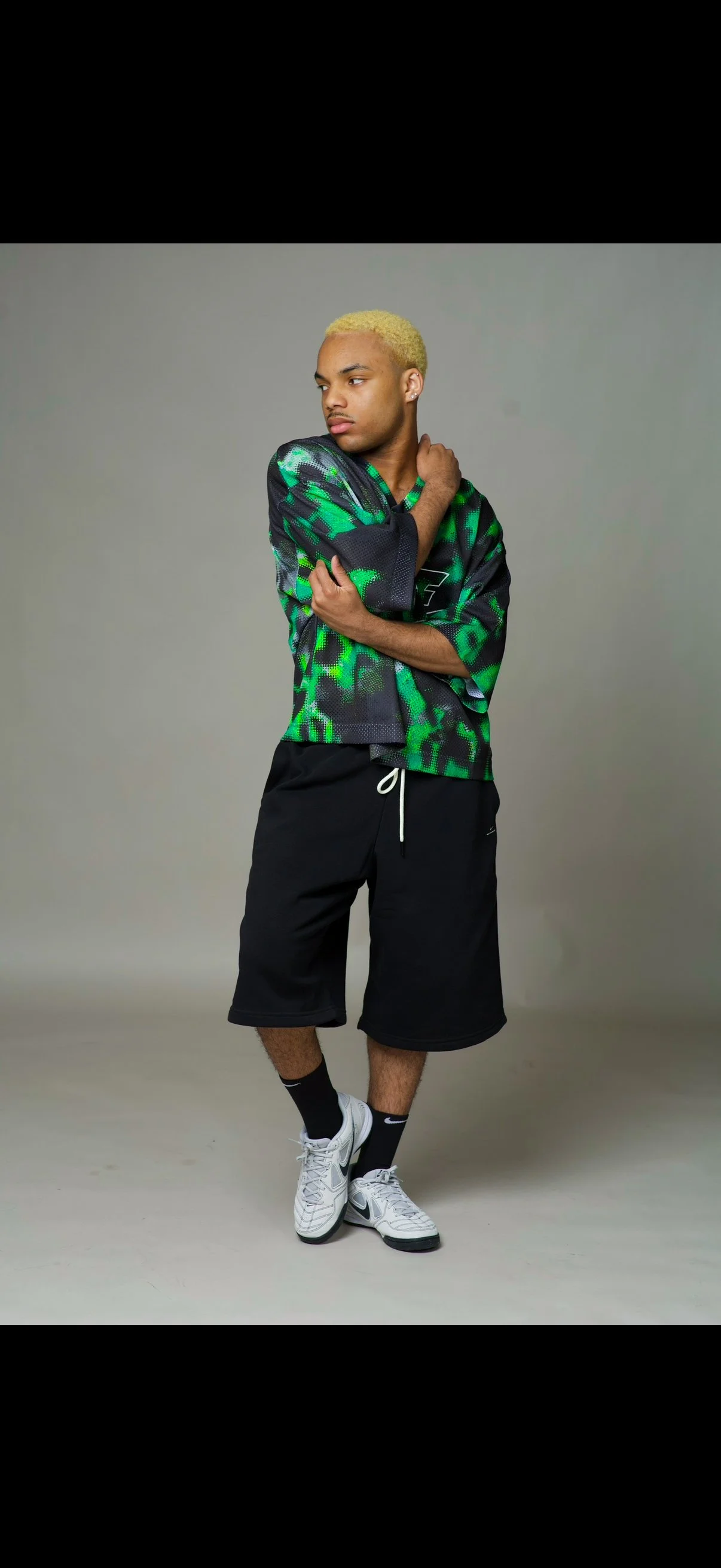 Young man with blond hair, wearing a green and black patterned sports jersey, black shorts, white athletic shoes, and black Nike socks, posed against a plain gray background.