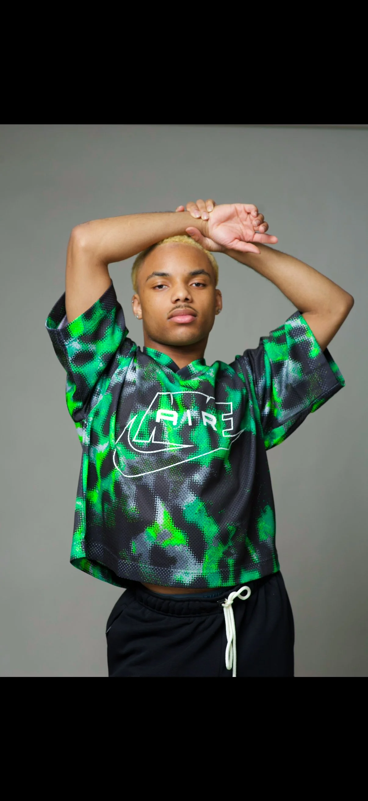 Young man with bleached blonde hair, wearing a black and green patterned sports jersey with the word 'PARIS' on it, and black shorts with a white drawstring. He stands against a plain gray background with one arm raised and resting on his head.