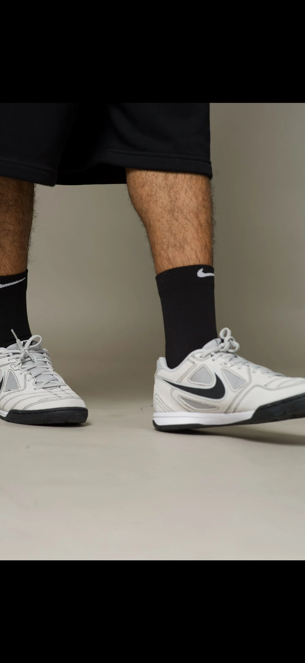 Close-up of a person wearing black Nike socks, white Nike sneakers with black swooshes, black shorts, and standing with one foot slightly in front of the other on a plain surface.