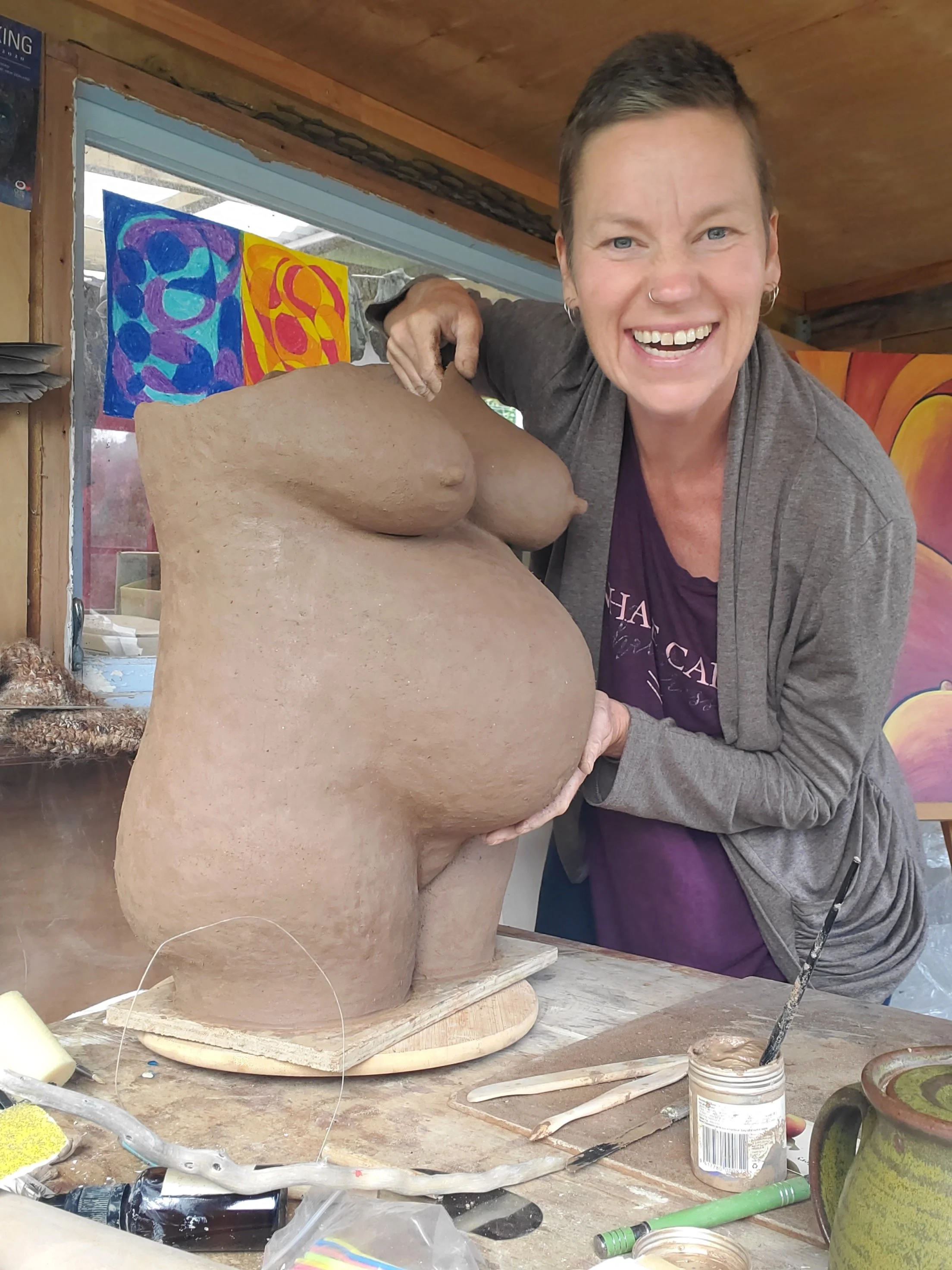 A woman smiling and working on a large clay sculpture in an art studio. The sculpture is of a pregnant torso. The background includes colorful abstract paintings and various art supplies on a wooden work surface.
