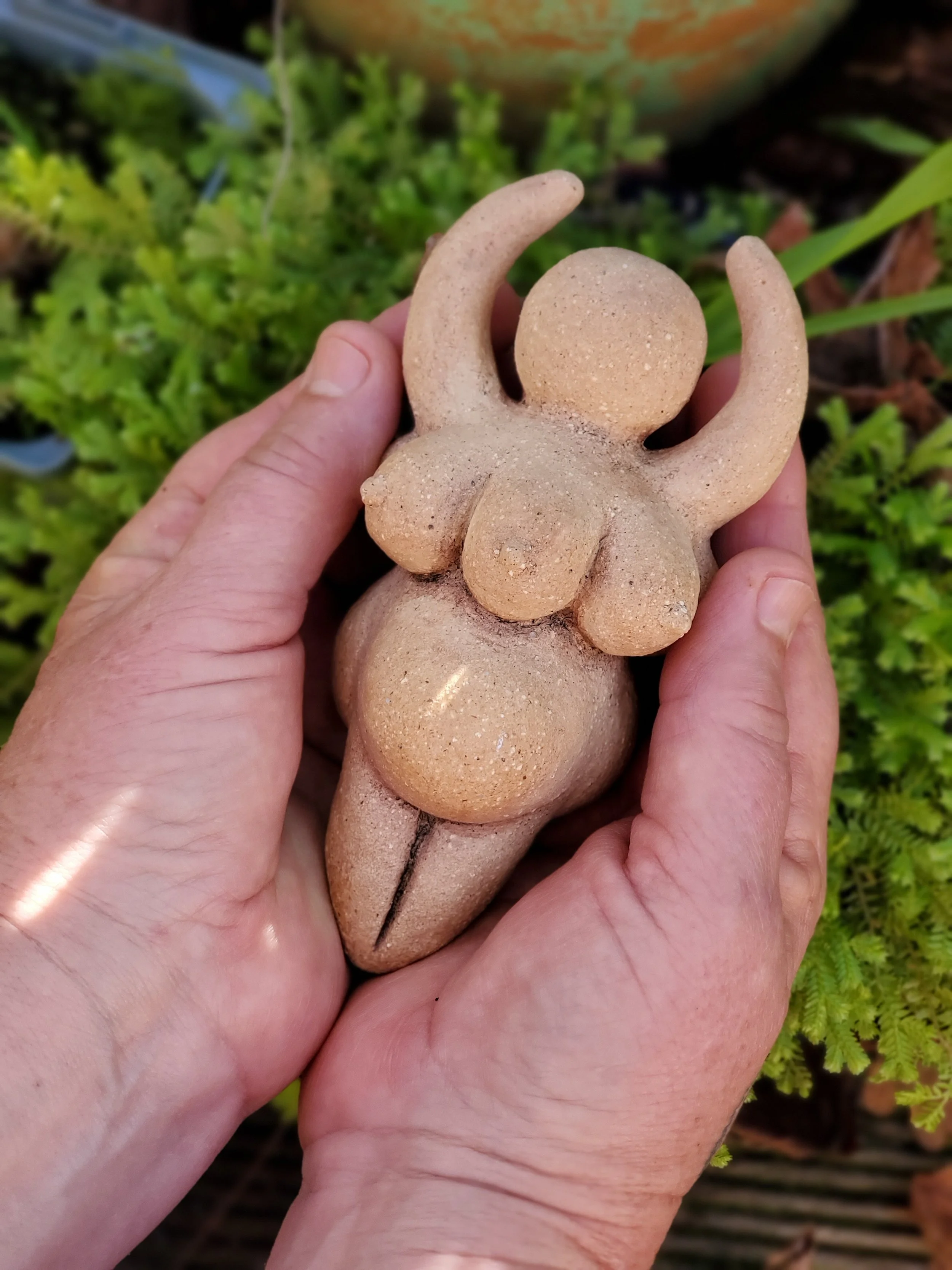 Hands holding a small pottery sculpture of a many breasted figure with arms raised, with greenery in the background.