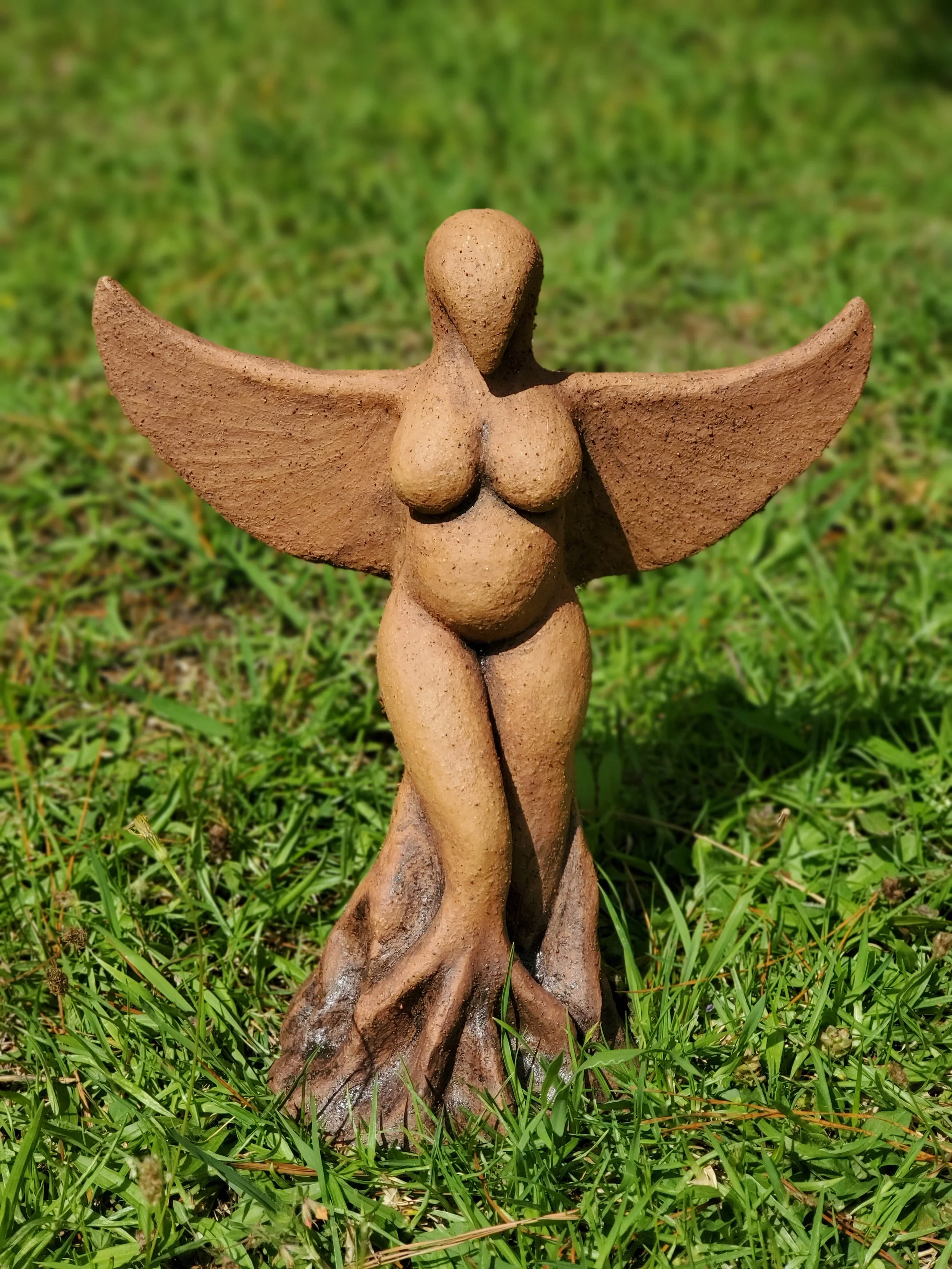 Clay sculpture of a feminine figure with wings, a birdlike head and roots, standing on a grassy surface.