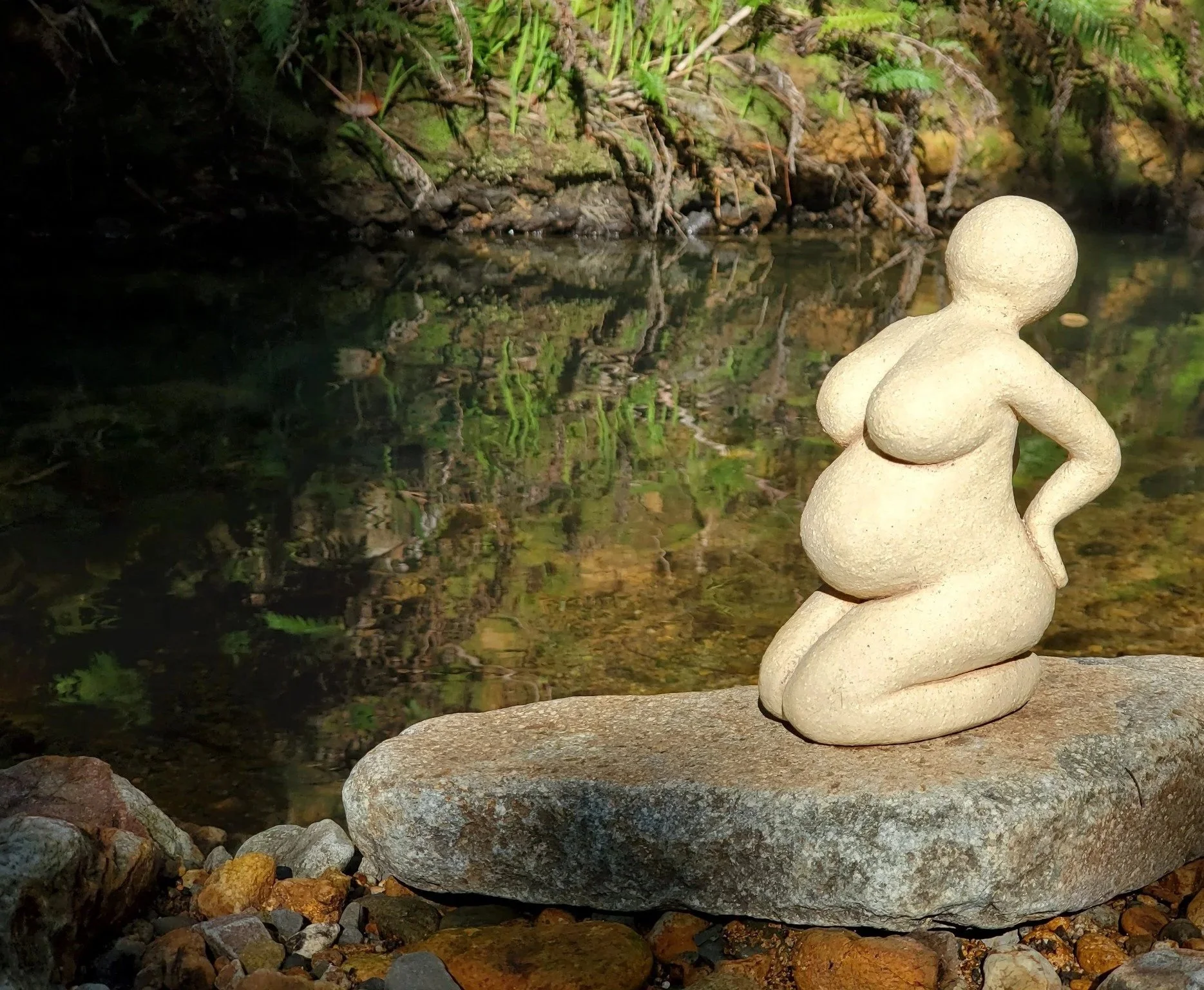 Pottery sculpture of pregnant woman on a rock in front of a river.