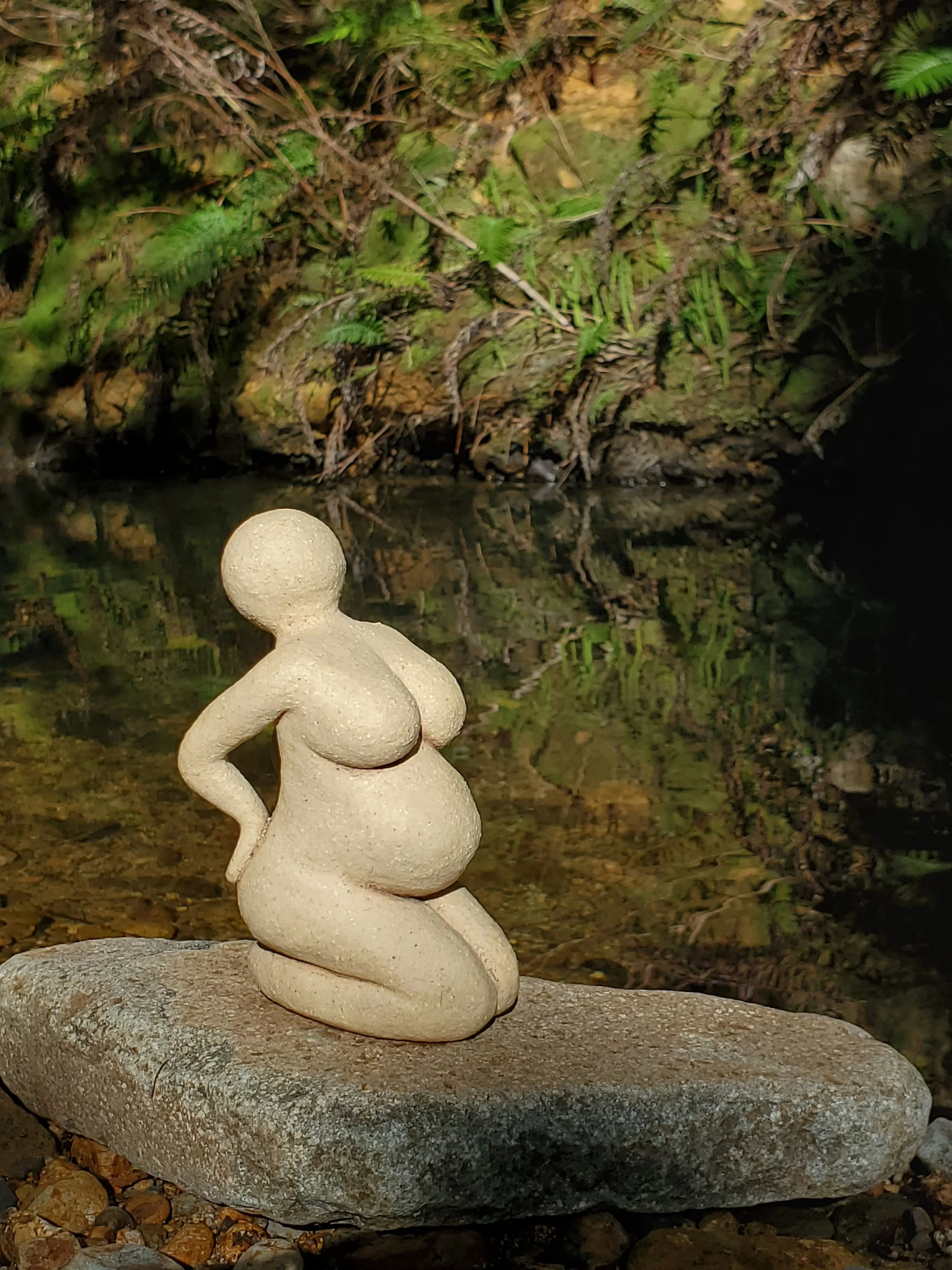 Pottery sculpture of pregnant woman on a rock in front of a river.