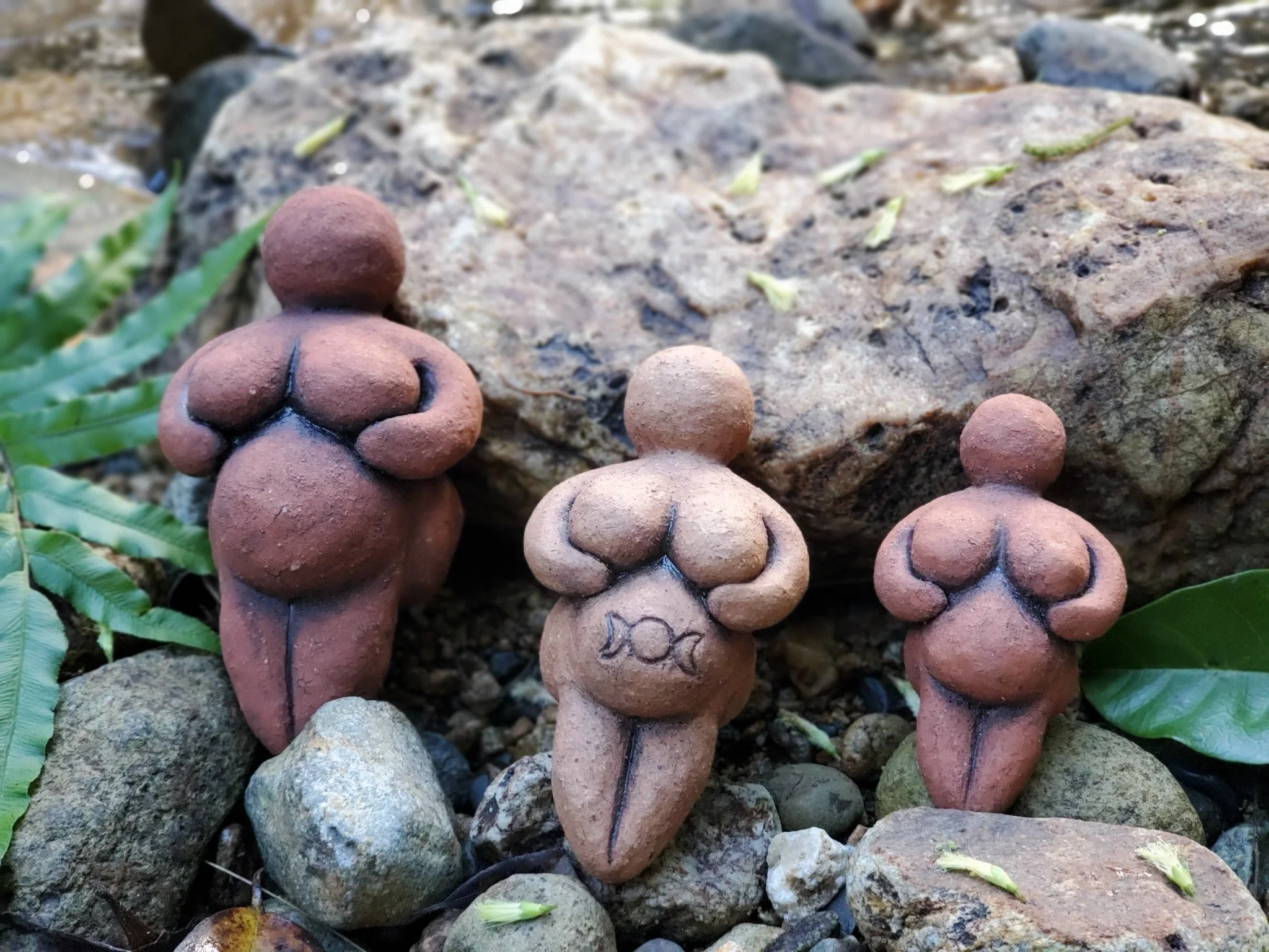 Small clay sculptures of women placed on rocks and leaves outdoors.