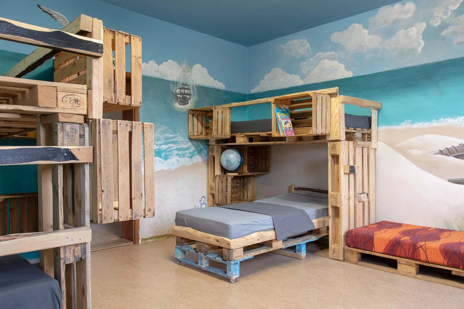 Ein Kinderzimmer mit Betten aus Holzpaletten, einer Wandmalerei mit Meeresmotiven und Strand, und verschiedenen pädagogischen Elementen. Ein Regal mit Büchern und eine Weltkarte sind ebenfalls sichtbar.