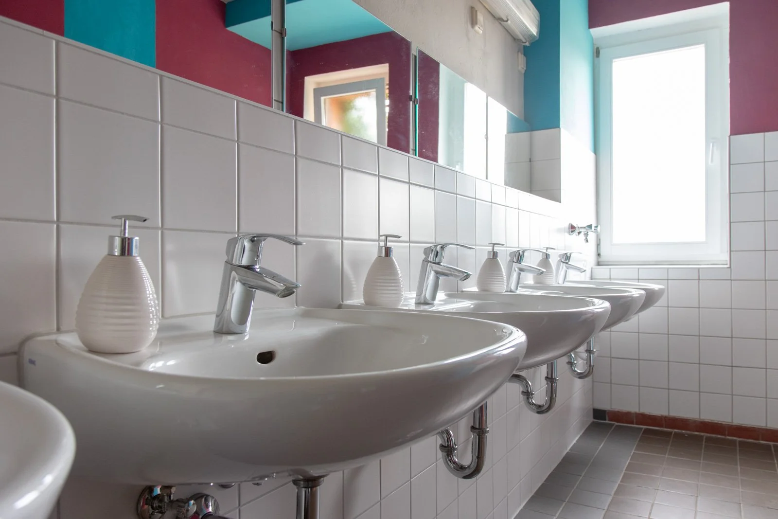 Mehrere weiße Waschbecken mit Wasserhähnen in einer öffentlichen Toilette, mit Seifenspendern davor und einem großen Fenster im Hintergrund.