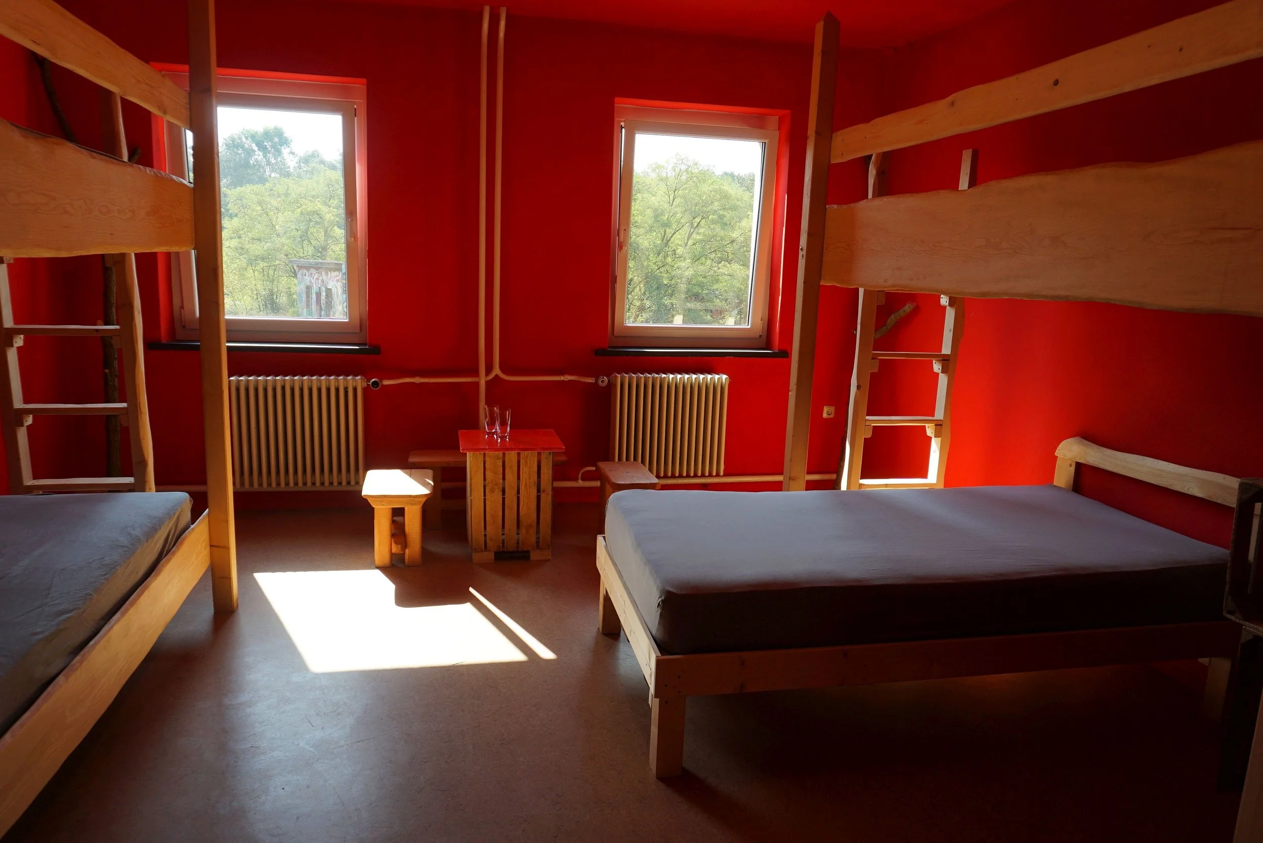 Ein Schlafzimmer mit roten Wänden und natürlichen Holzmöbeln, inklusive zwei Etagenbetten, Fenster mit Blick auf Bäume, Heizkörper, und kleine Lampen.