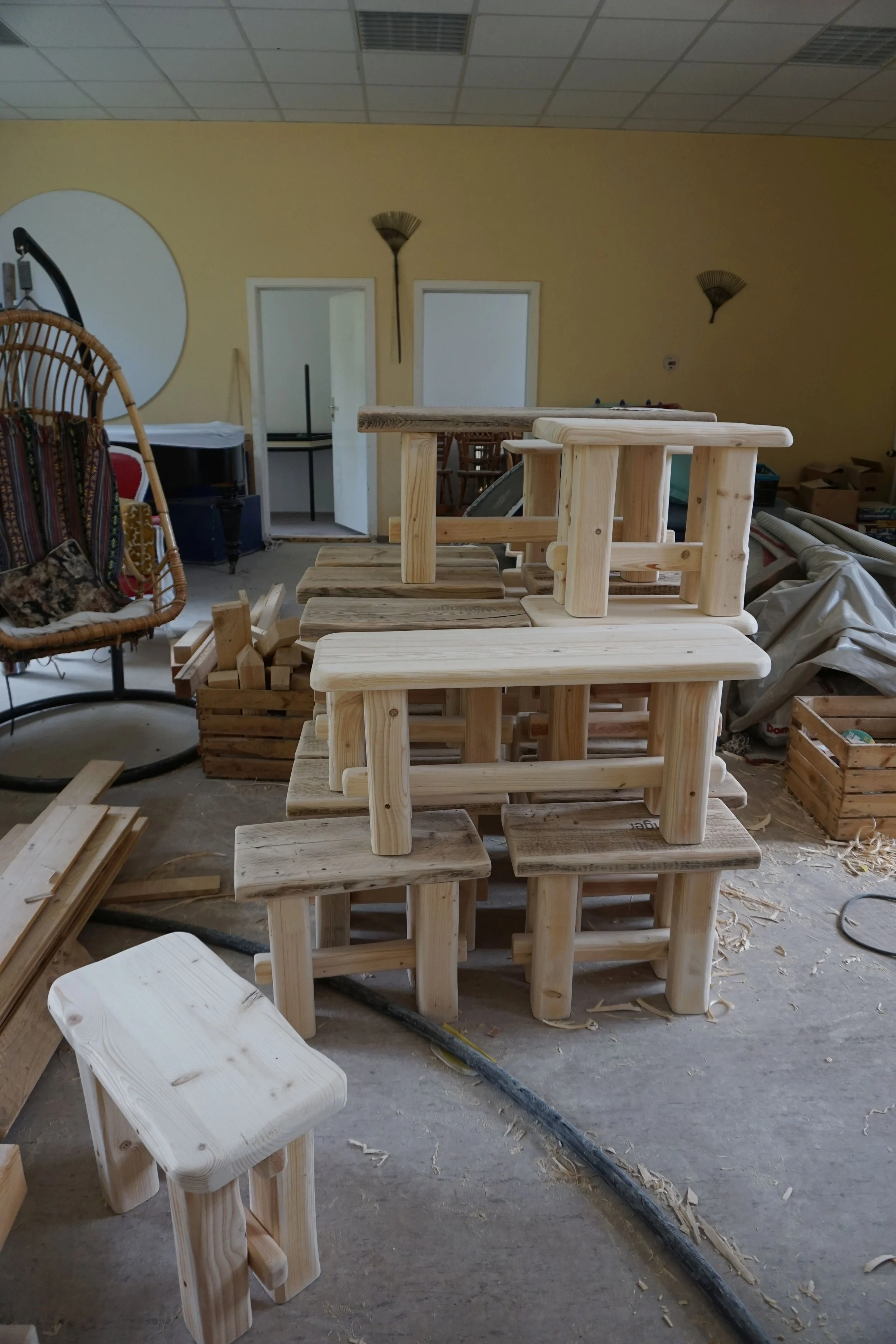 Holzstühle in einem Raum, die noch montiert werden. Im Hintergrund sind weitere Möbel, eine Lampe und Papiersäcke sichtbar.