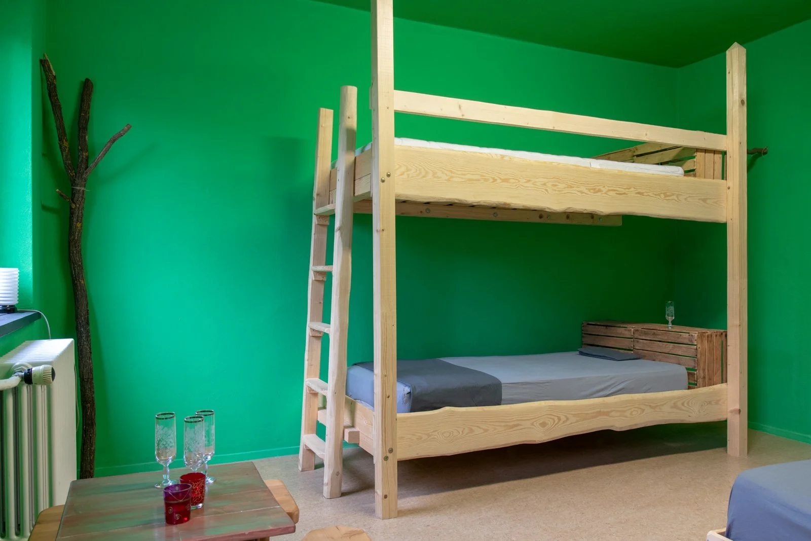 Ein Zimmer mit grünen Wänden, einem Etagenbett aus Holz, einem kleinen Tisch mit Gläsern, einem Fenster, einem Heizkörper und einer Lampe auf einem Nachttisch.