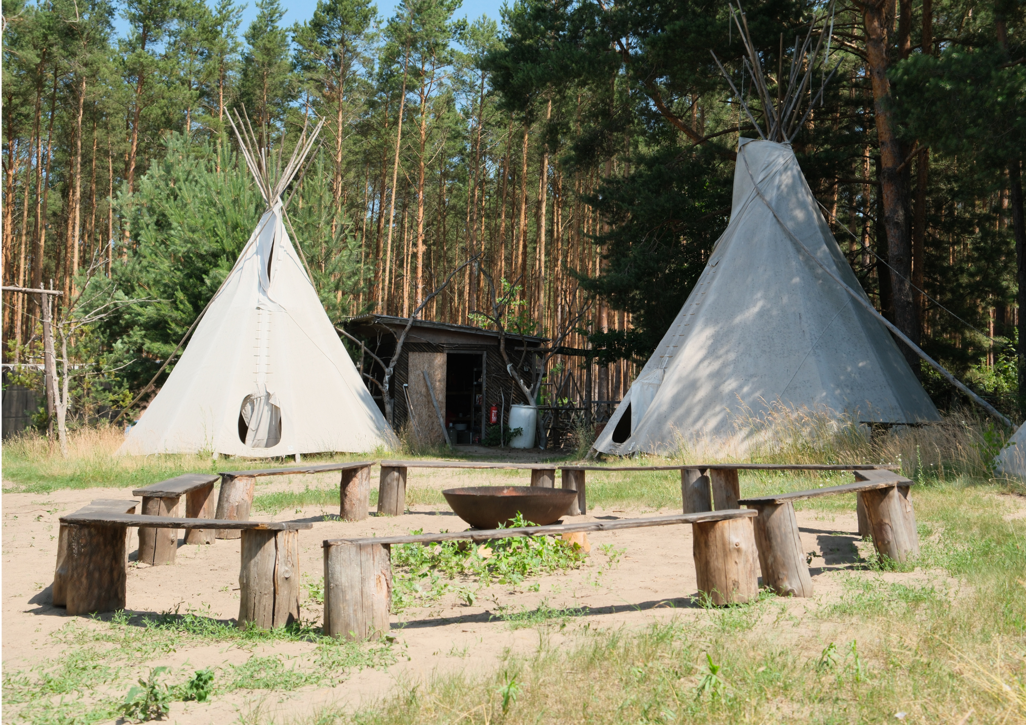 Zwei Tipis in einem Waldcamp, umgeben von Bäumen, mit Holzstuhl-Reihen im Vordergrund.