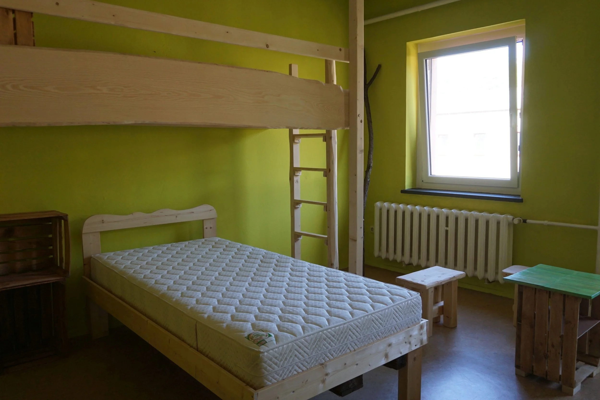 Ein Schlafzimmer mit grünen Wänden, einem Fenster, einem Bett mit Matratze, einem kleinen Tisch aus Holz, einer Holzwand mit Leiter, Radiator unter dem Fenster, Holzdekor, natürliche Beleuchtung.