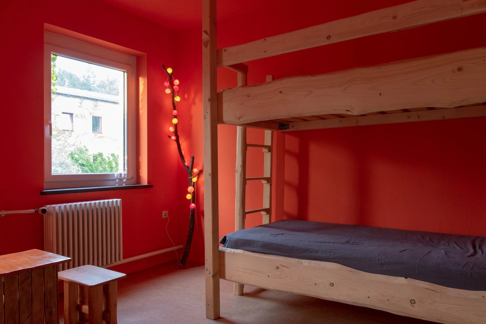 Ein Kinderzimmer mit orangefarbenen Wänden, einem Fenster, einer Heizung, einem kleinen Tisch und Stühlen, einem vertikalen Ast mit bunten Lichterketten und einem klassischen Bett mit einer oberen und unteren Liegefläche aus Holz.