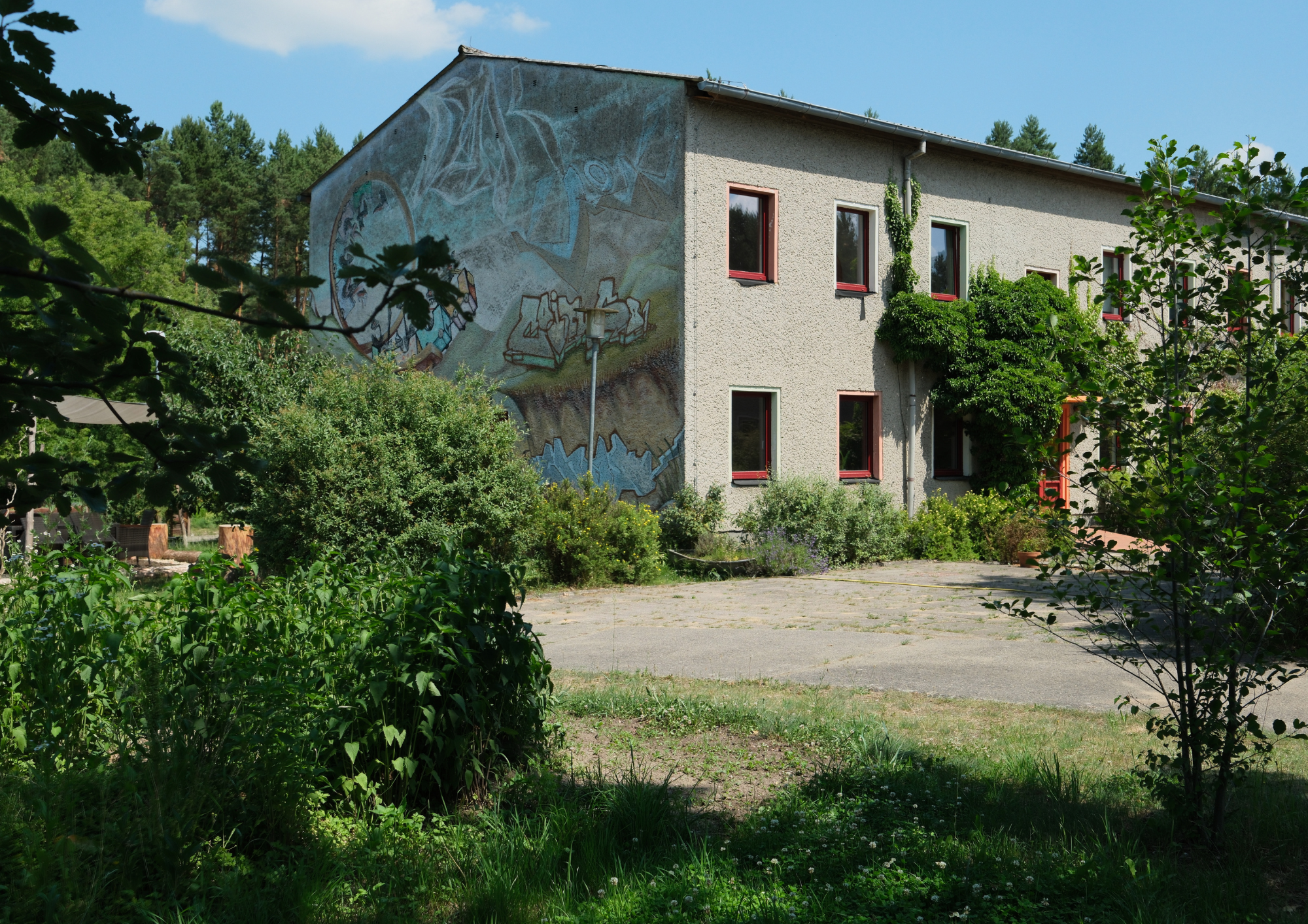 Ein mehrstöckiges Gebäude mit einem Wandgemälde auf der linken Seite, umgeben von Büschen und Bäumen, auf einer gepflasterten Fläche bei sonnigem Wetter.