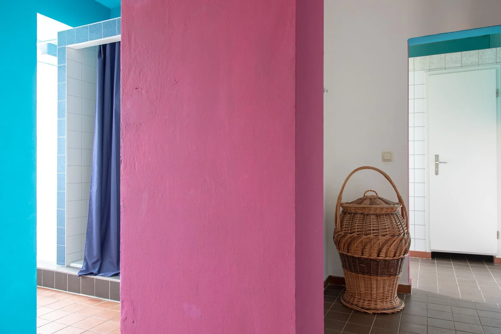 Badezimmer mit pinkener Wand, Wäschekorb, Türen zu duschraum mit blauer Duschvorhang und einem hellblauen Türrahmen.