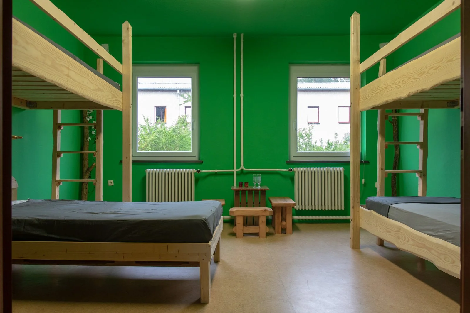 Ein Zimmer mit grünen Wänden, zwei Etagenbetten aus Holz, zwei Fenster mit Ausblick auf Bäume, Heizkörper und ein kleines Tischchen mit zwei Gläsern