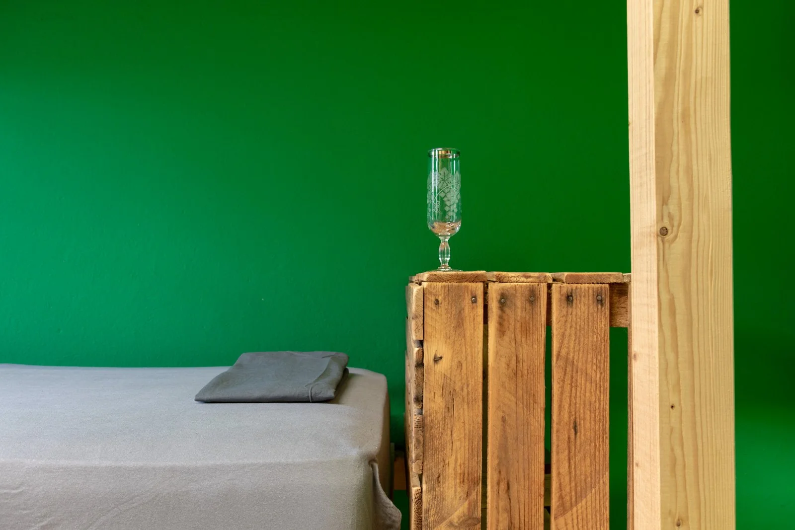 Ein Schlafzimmer mit grüner Wand, ein Bett mit grauem Kissen, eine Holz-Paletten-Nachtkästchen, eine leere Sekt- oder Weinglas auf dem Nachttisch.