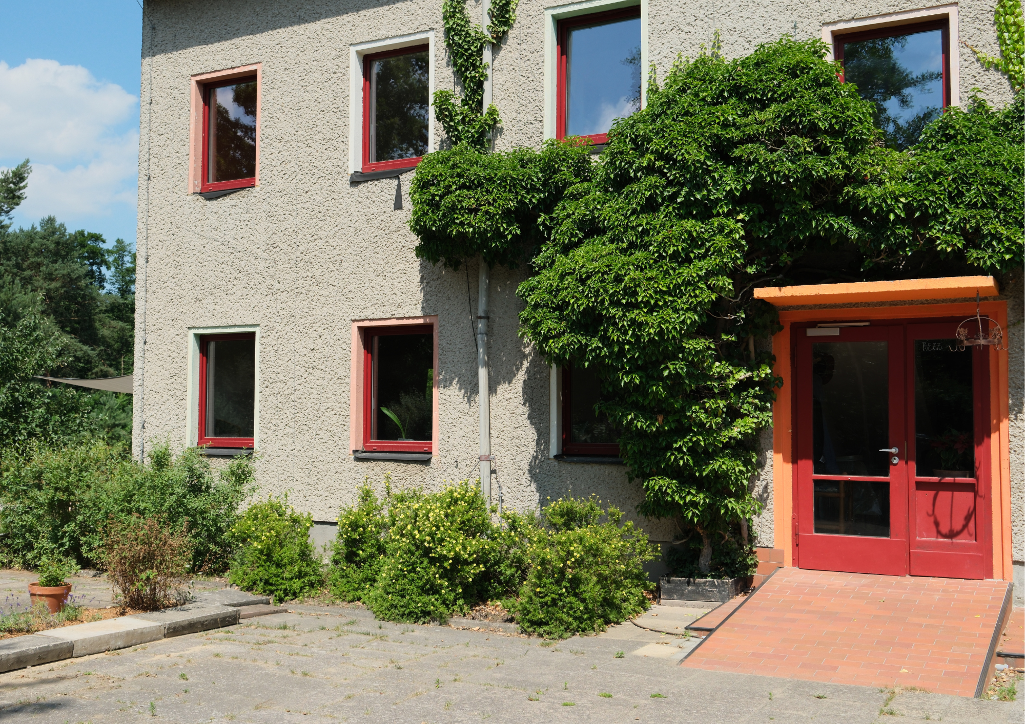 Das Bild zeigt eine zweistöckige Hausfassade mit einer Raufassade und mehreren Fenstern mit roten Rahmen. Es gibt einen Eingang mit einem roten Tür mit Glasfenster, über dem eine Überdachung mit leuchtender orangefarbener Umrahmung. Das Haus ist von 