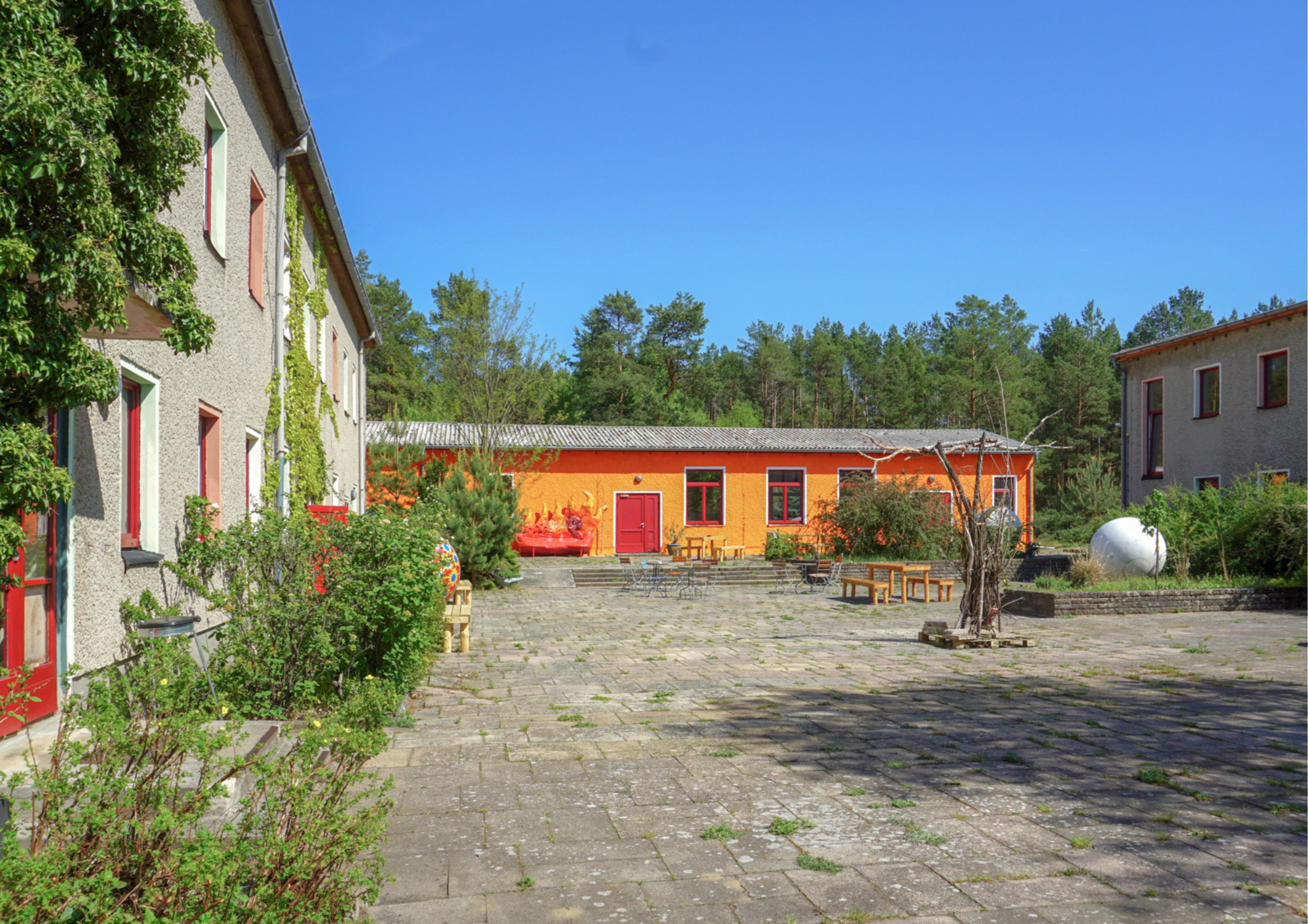 Ein Innenhof mit gepflastertem Boden, umgeben von mehrstöckigen Gebäuden mit Fenster und Türen, einige mit roten Rahmen. Im Hof sind Gartenmöbel, Pflanzen und dekorative Gegenstände, mit Bäumen im Hintergrund unter blauem Himmel.