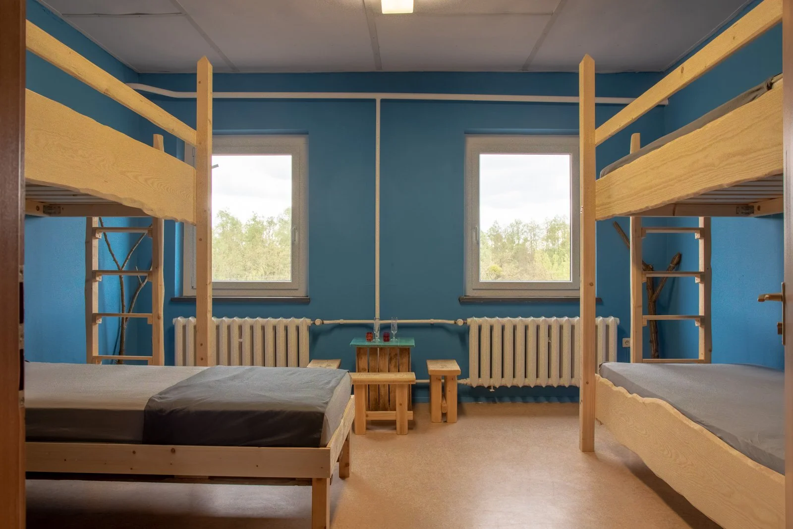 Ein Schlafzimmer mit blauer Wand, zwei Fenster, zwei Etagenbetten aus Holz, eine kleine mit zwei Hockern und ein Tisch in der Mitte, Heizkörper unter den Fenstern.