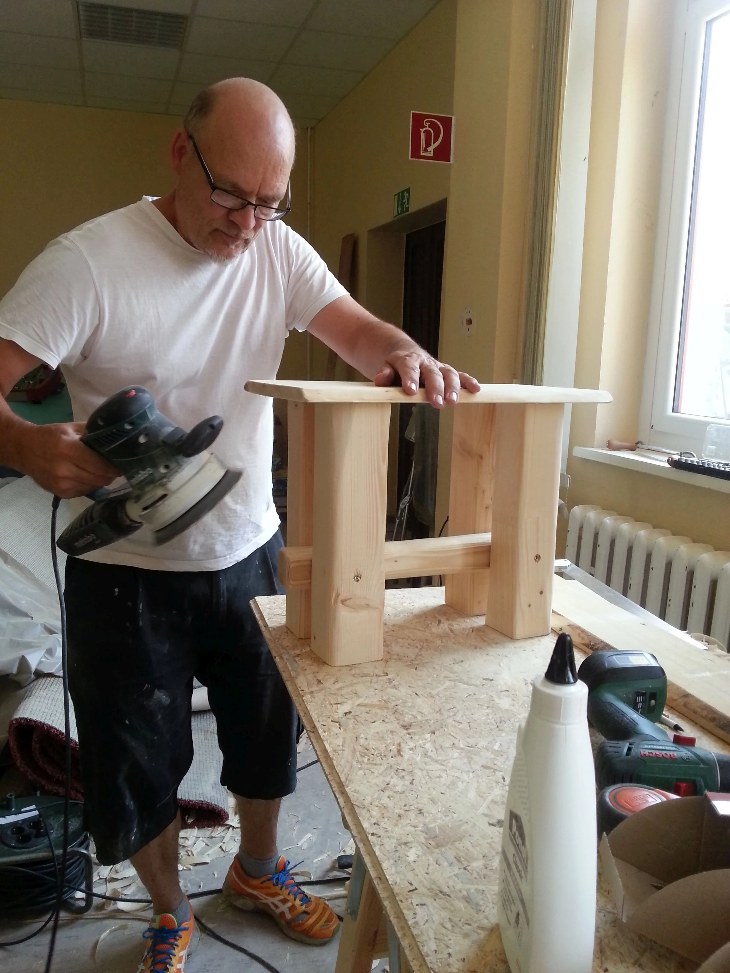Ein Mann bearbeitet Holz mit einer Stichsäge in einem Workshop, er trägt eine weiße T-Shirt, schwarze Shorts und blaue Schuhe.