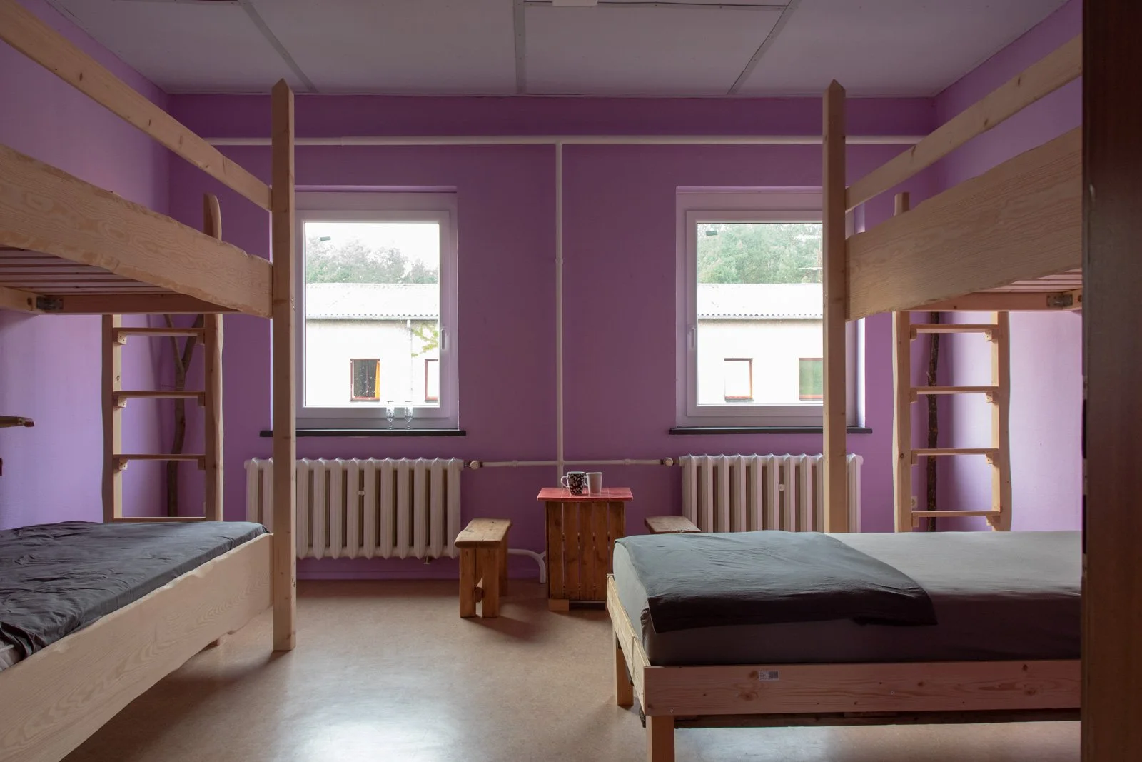 Ein Schlafzimmer mit zwei Hochbetten mit Geländern auf beiden Seiten, zwei Betten jeweils mit dunklem Bezug, ein kleines Holzregal in der Mitte des Raumes mit Kaffeetassen, zwei Fenster an der Wand, eine Heizung unter den Fenstern, die Wände sind lil