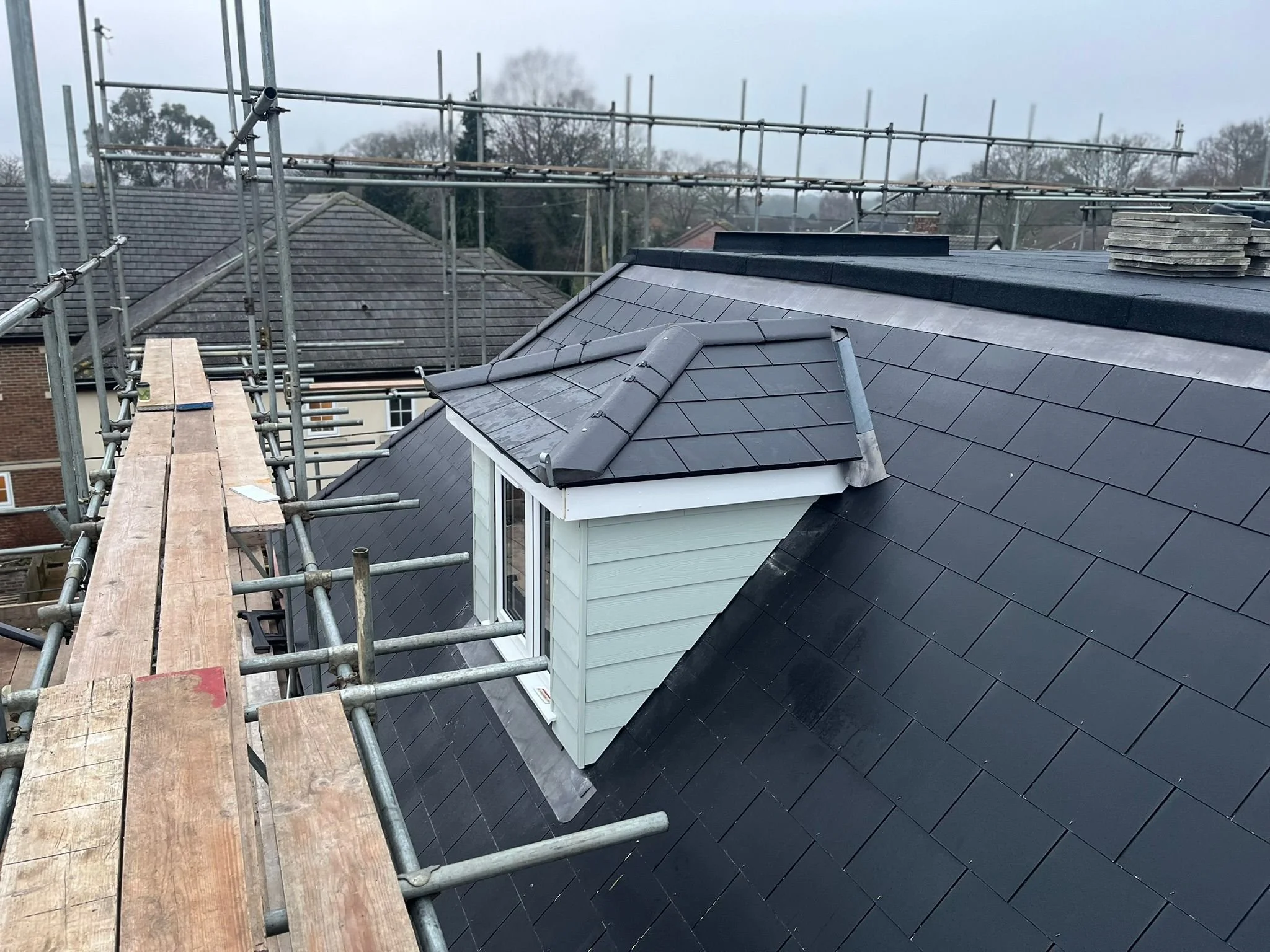 Construction scaffolding on a roof with a small dormer window, shingles, and a stack of tiles, overcast sky in the background.