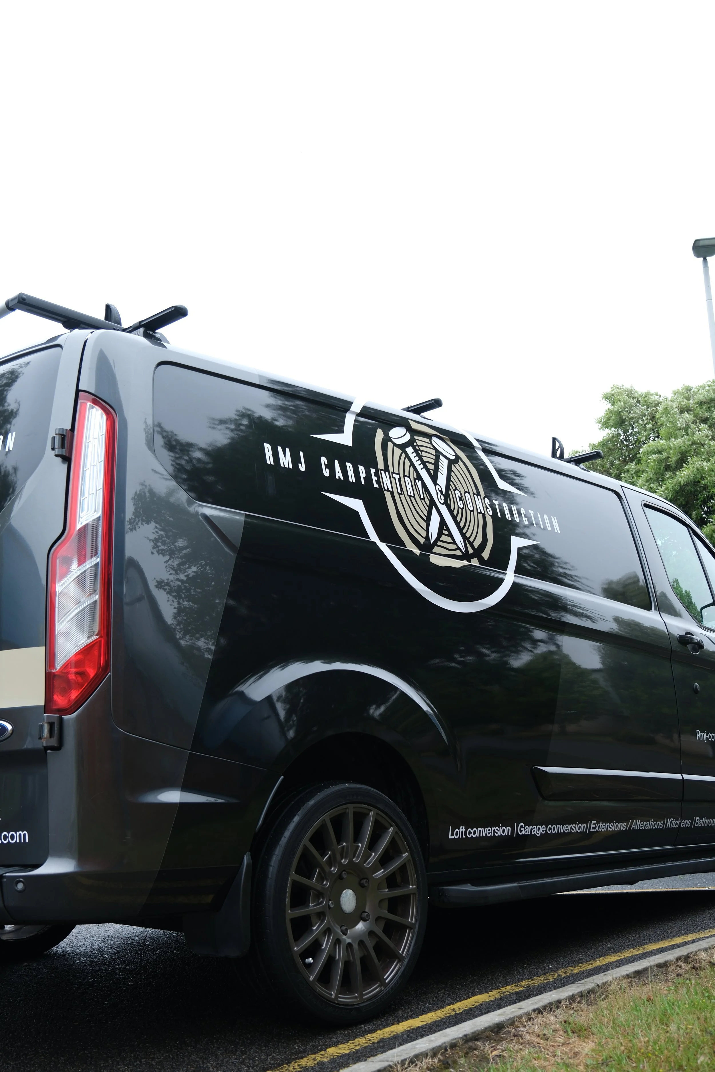 A black van with the logo and text 'RMJ Carpenter & Construction' on its side, parked in a lot. The van features a graphic of two crossed nails with a wood grain background and text listing various home renovation services.