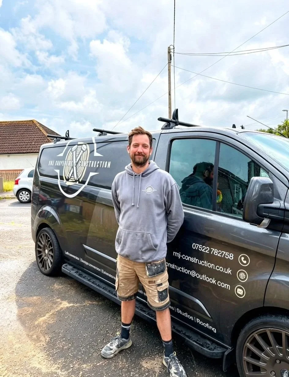 A man standing in front of a black company van with construction branding, wearing a gray hoodie, tan shorts, and sneakers, smiling and posing outdoors under a partly cloudy sky.