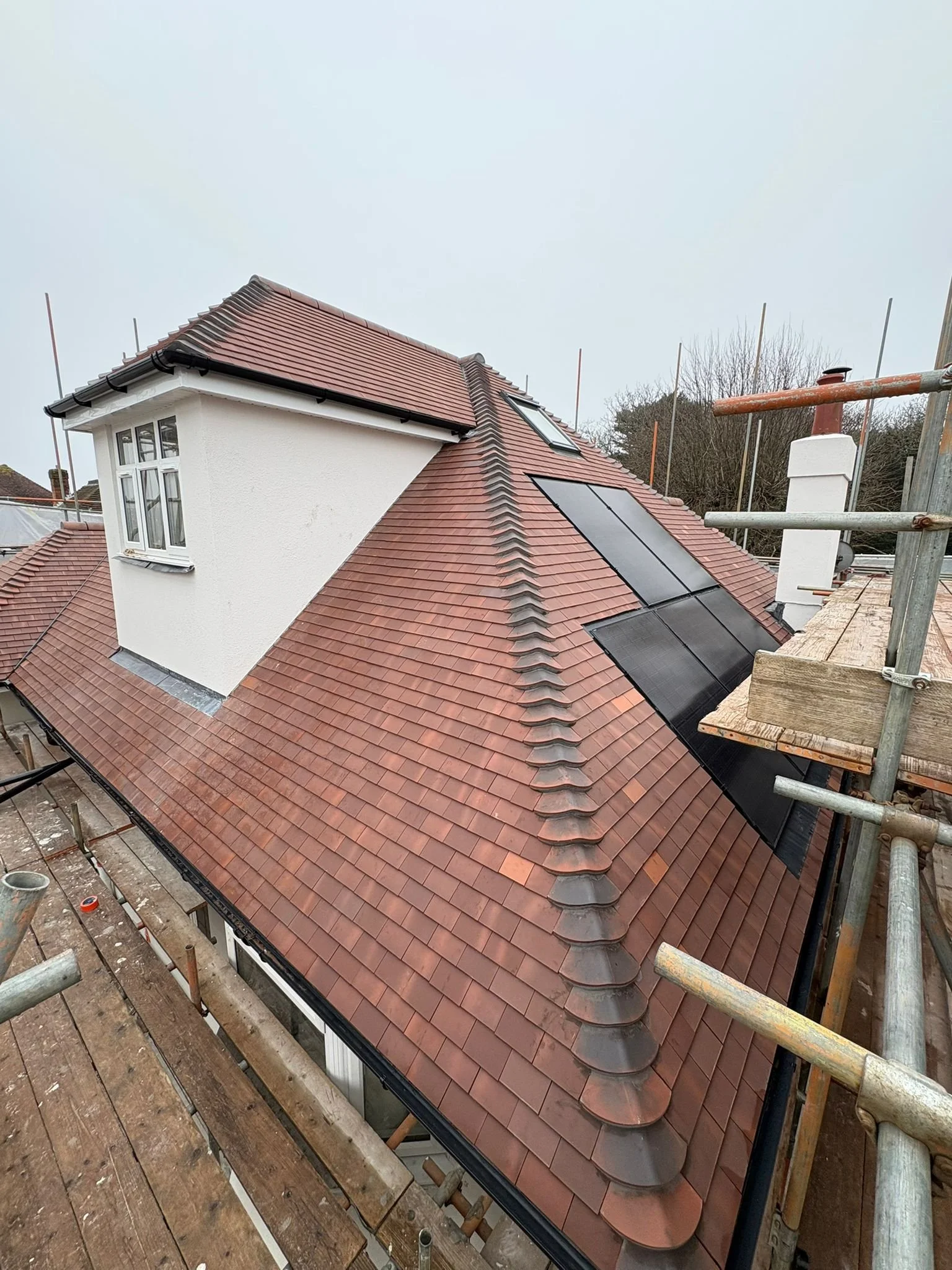 A house roof under construction with red clay tiles, a small dormer window, and solar panels, surrounded by scaffolding.