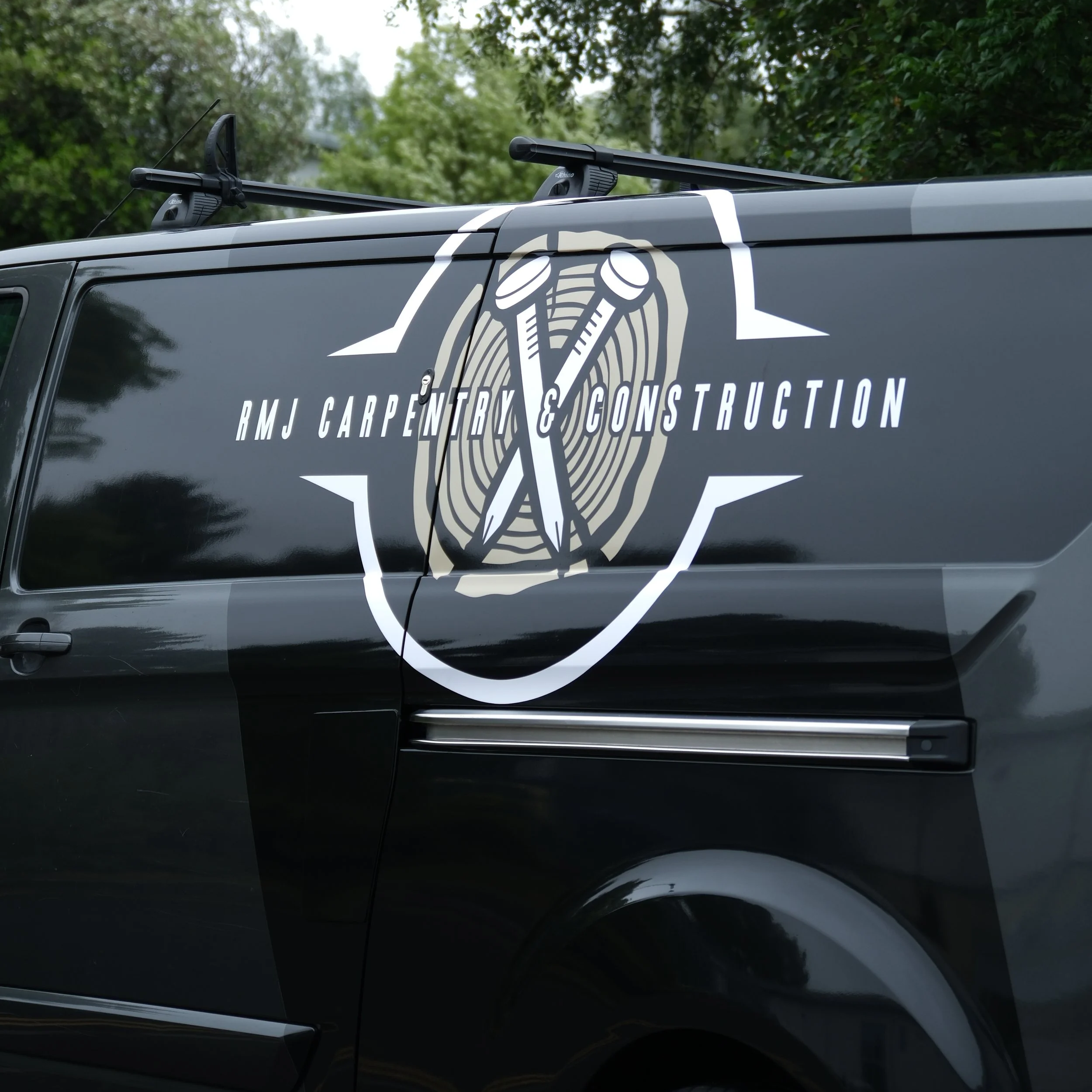 Side of a black vehicle with a logo and business name 'RMJ Carpentry & Construction' and a roof rack with crossbars. The background shows trees and the sky.
