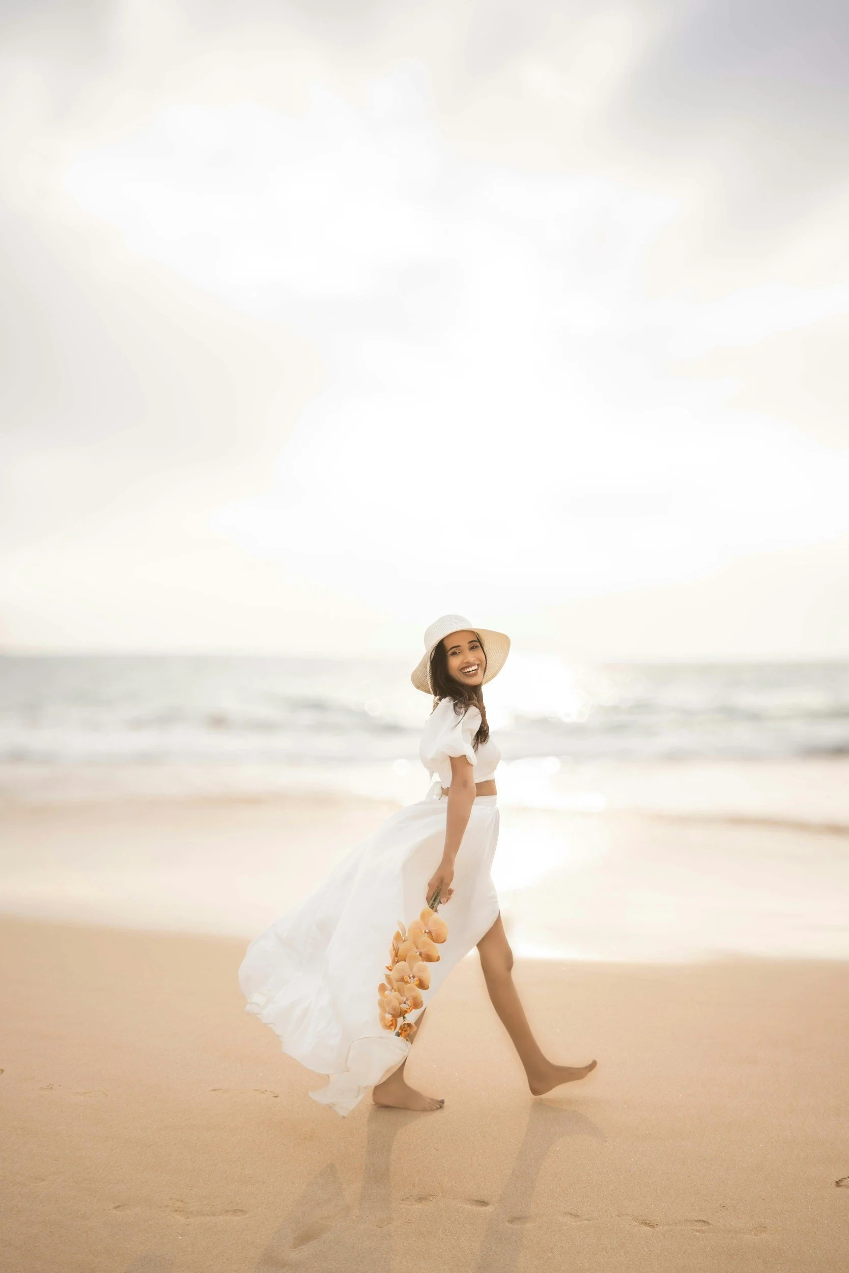 A woman in a white dress and wide-brimmed hat walking barefoot on a sandy beach, holding a bouquet of peach-colored flowers, with the ocean and cloudy sky in the background.